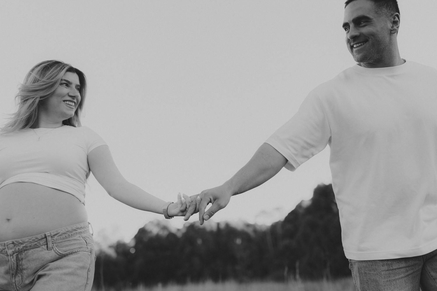 Pregnant couple holding hands smiling in field, black and white maternity photo.