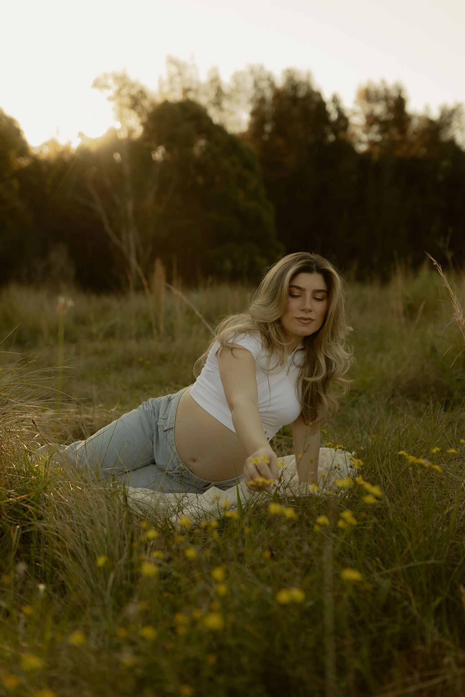 Pregnant woman in white crop top and jeans sitting in golden field at sunset surrounded by wildflowers.