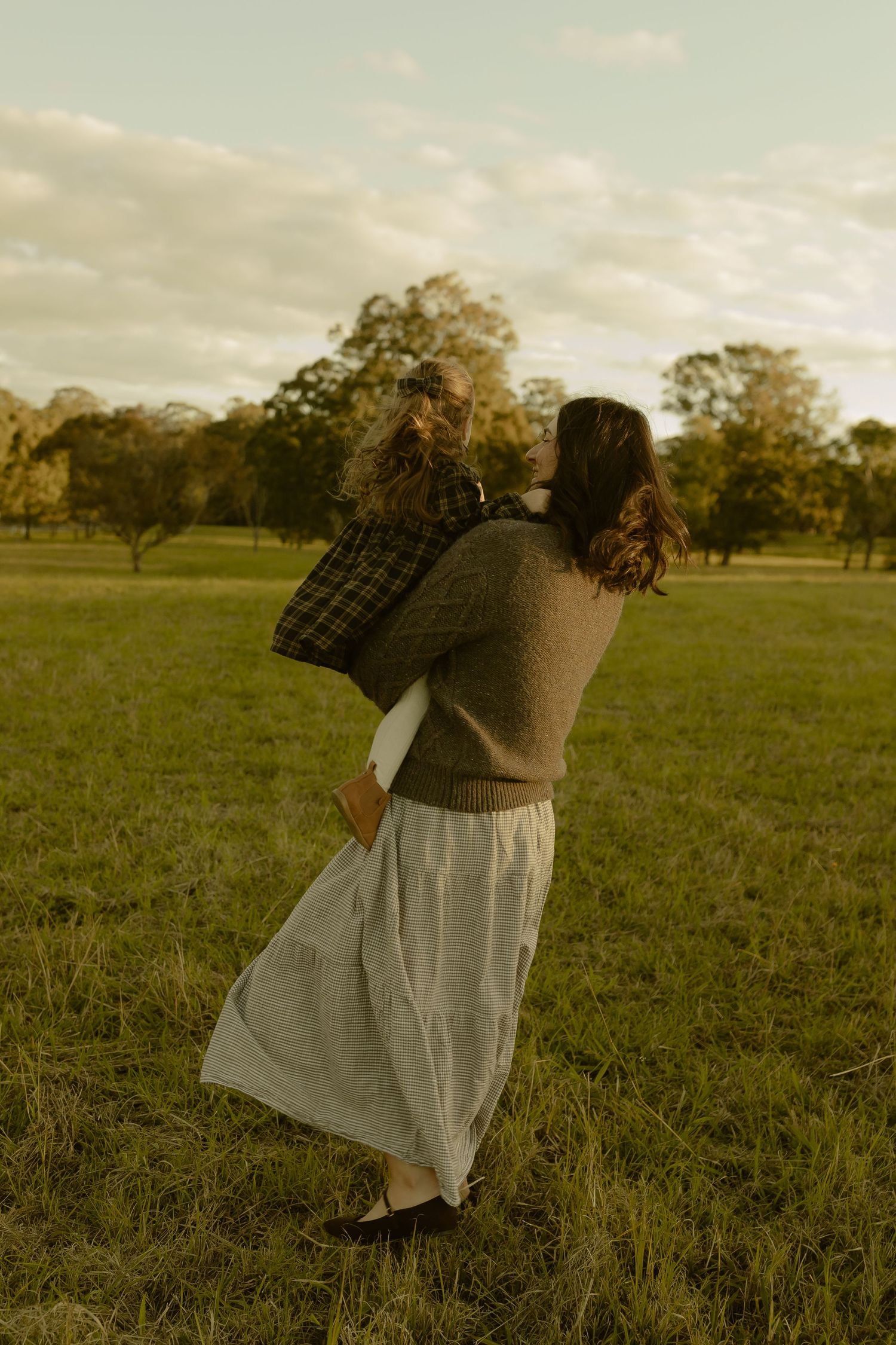 Woman in beige sweater and white skirt carries child across lush green park field at golden hour.