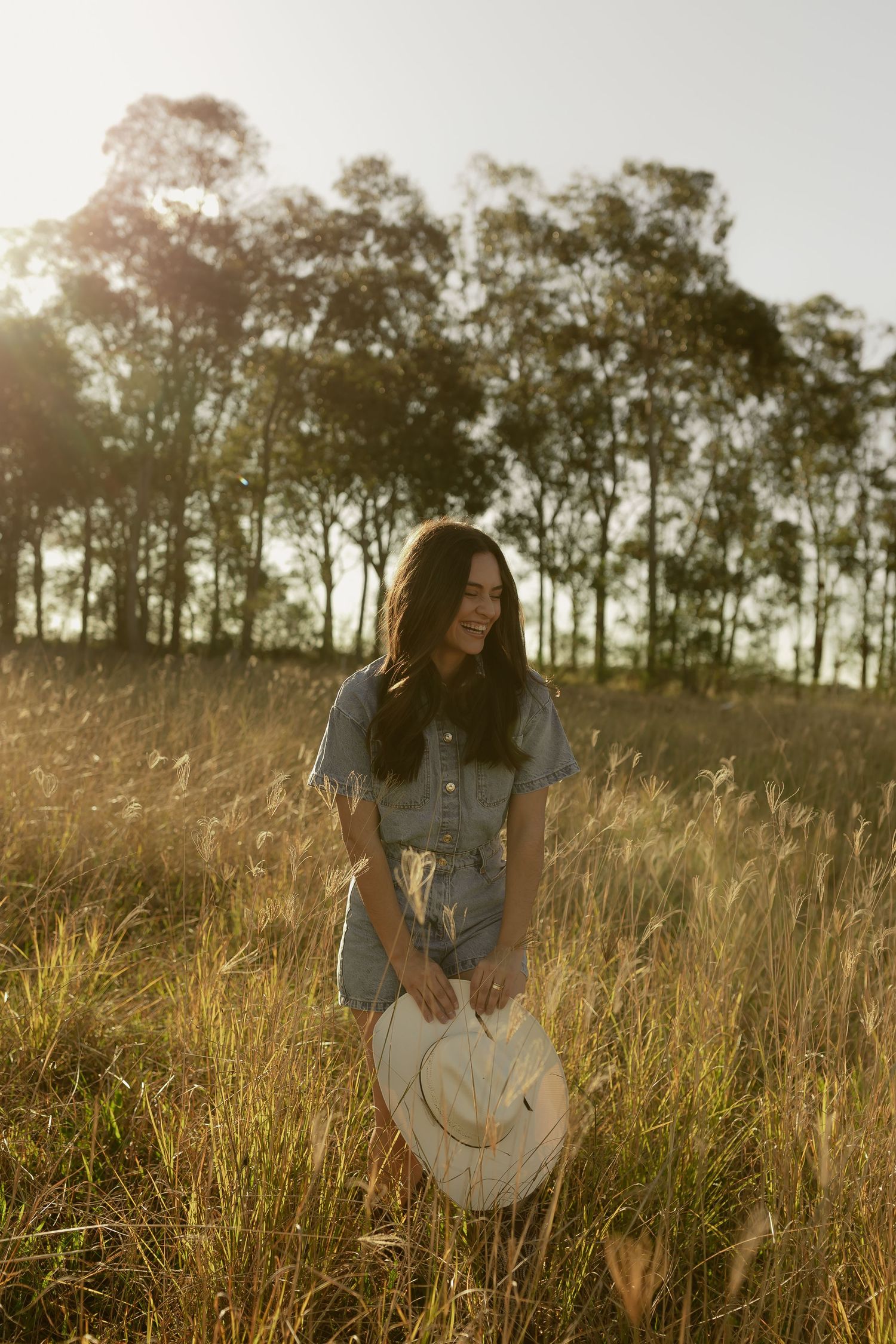 Young woman in denim romper holding straw hat standing in golden grass field with trees at sunset.