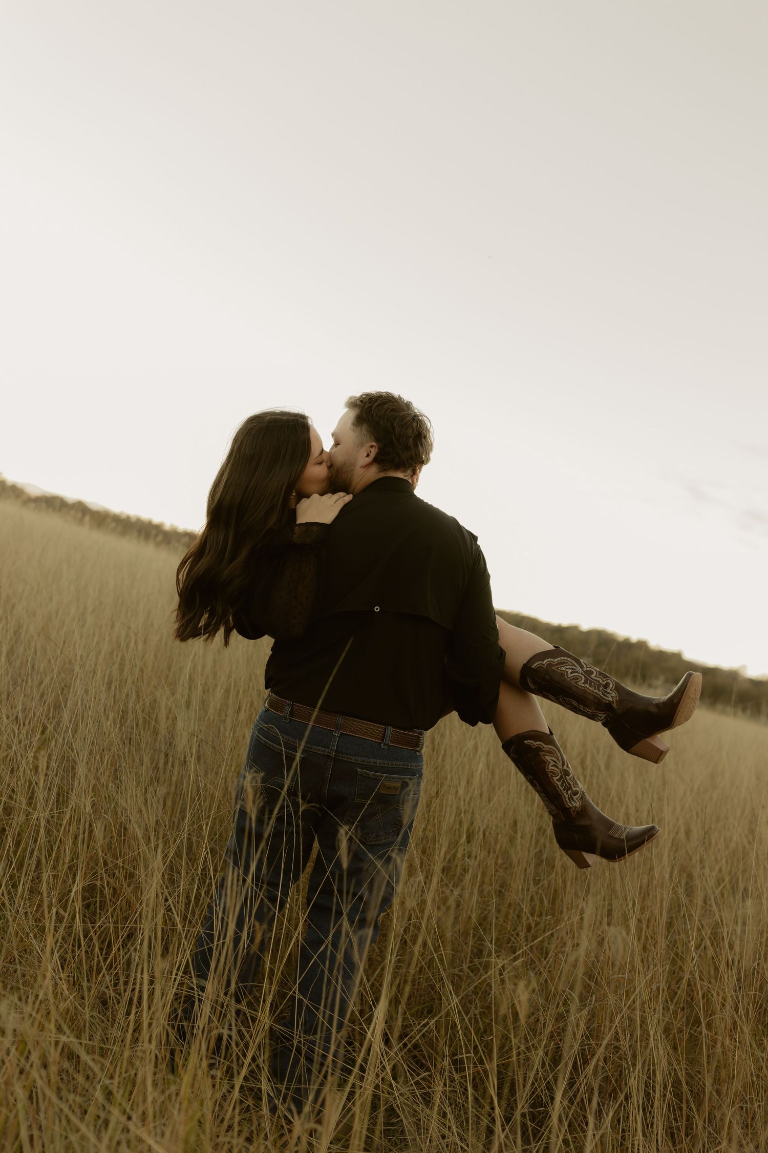 Romantic couple kissing in golden field, man lifting woman wearing cowboy boots at sunset.