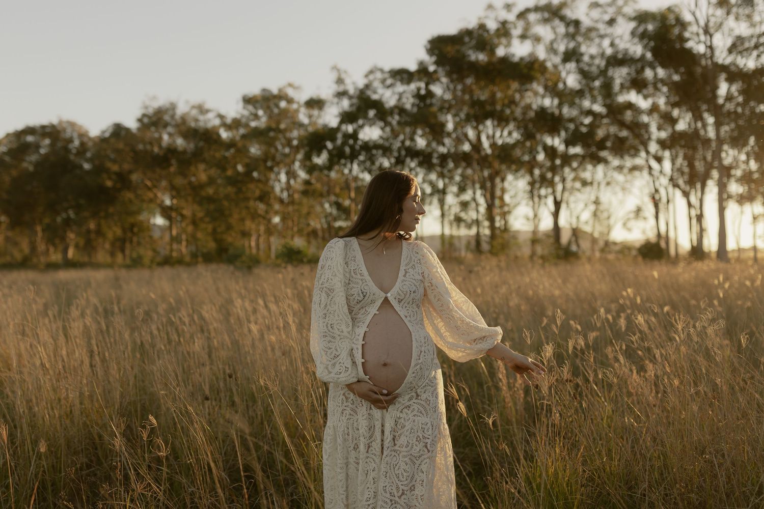 Pregnant woman in white lace dress walking through tall grass field at sunset.