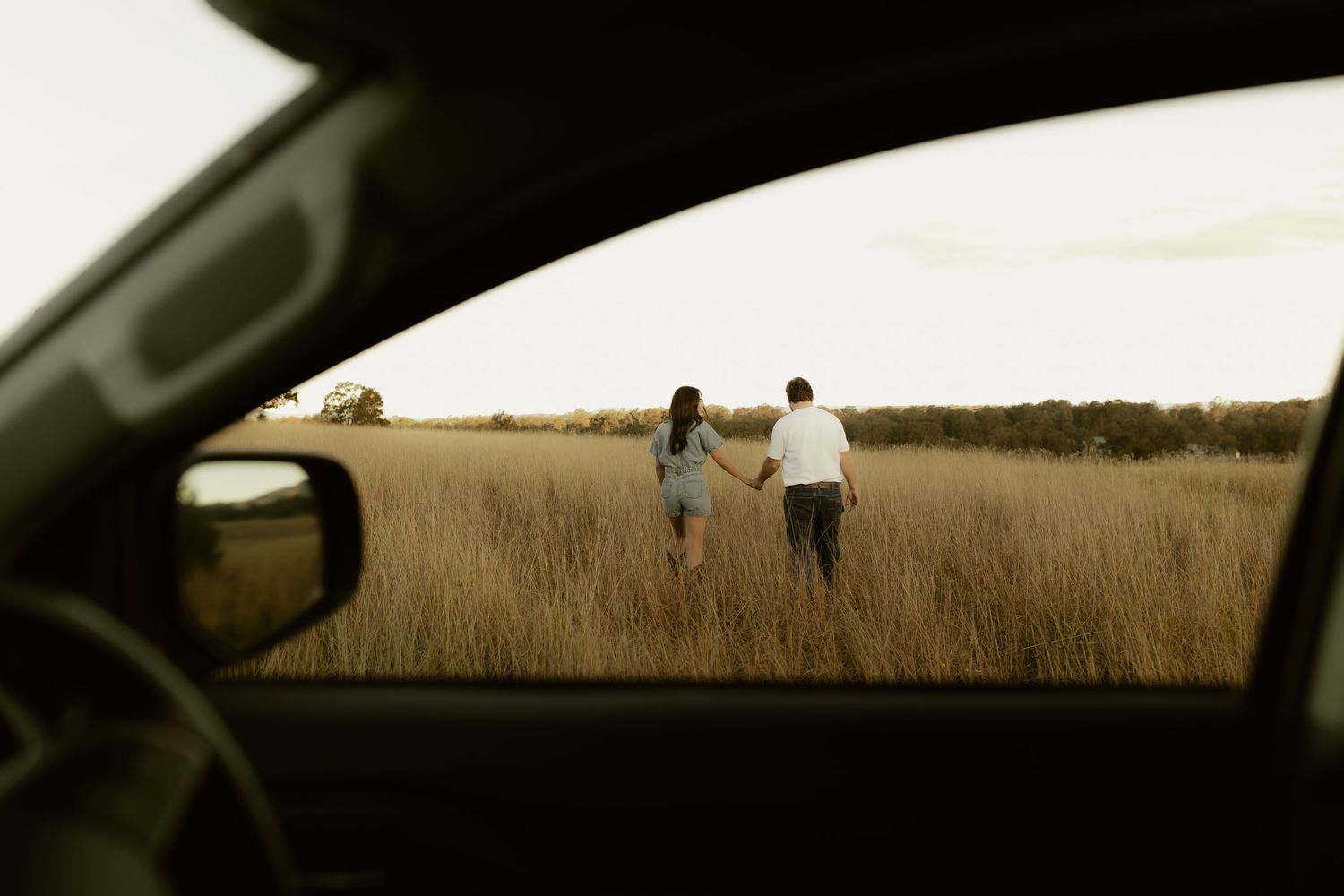 A couple holds hands walking through a golden grass field, viewed through a car window.