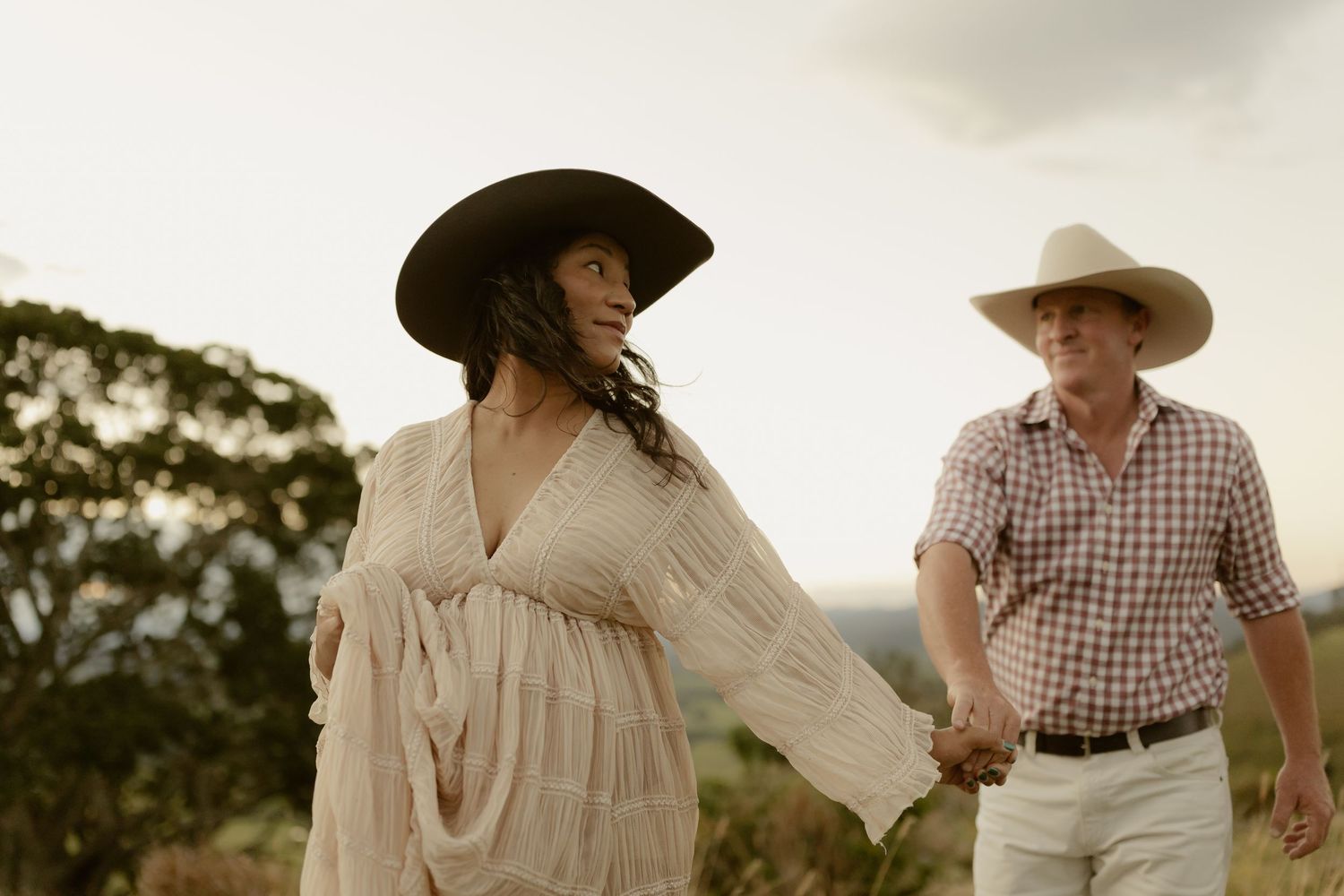 Couple in cowboy hats holding hands outdoors, woman in white dress, man in plaid shirt, romantic countryside setting.