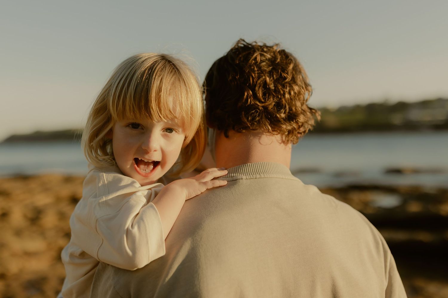 Laughing blonde toddler being held by a curly-haired parent on a sunny beach.