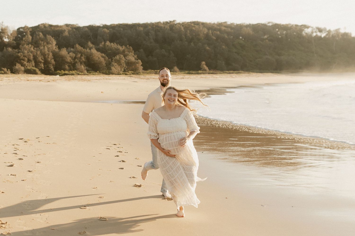 Couple in white outfits running joyfully on a sunny beach, waves and golden sand in the background.