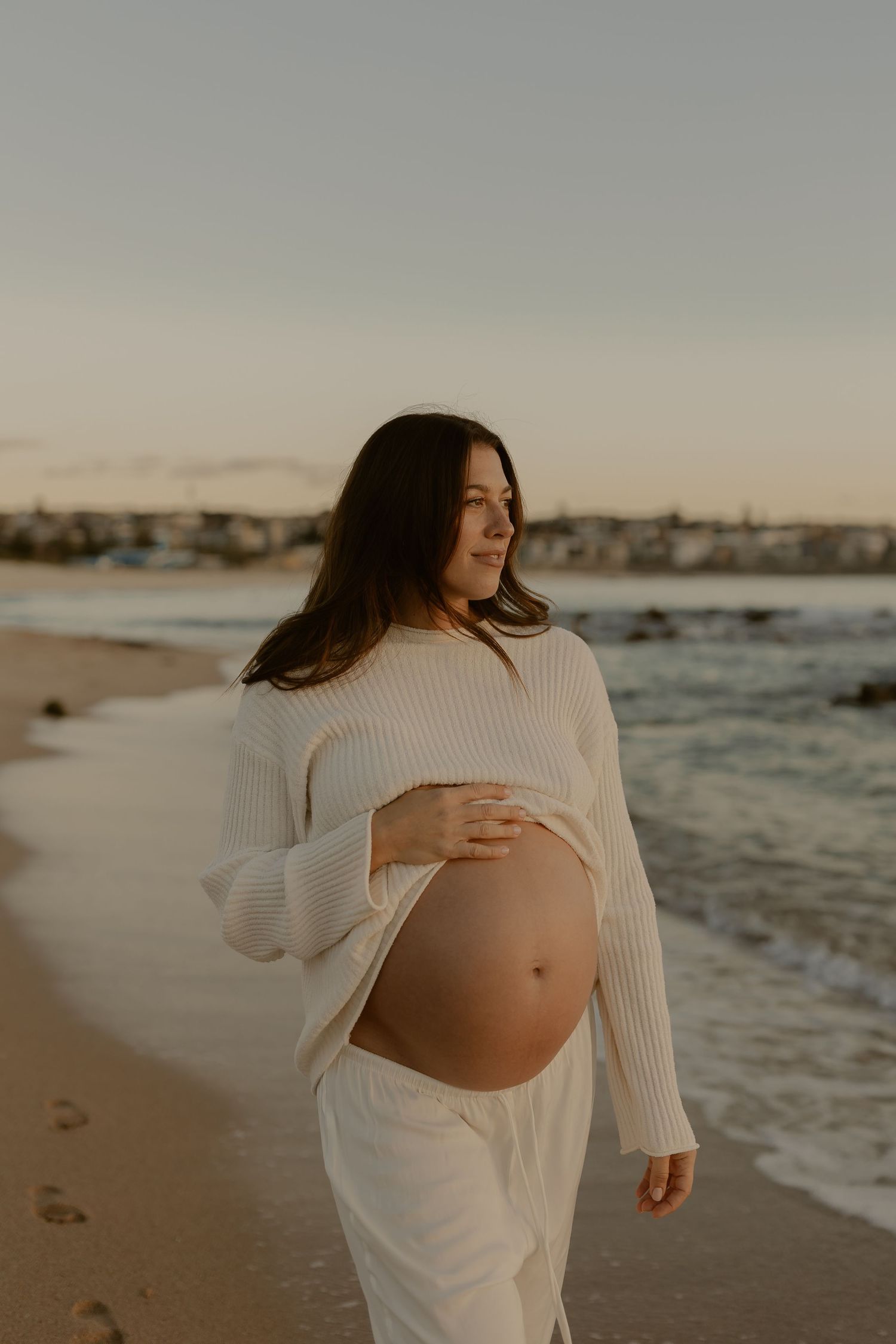 An expectant mother in white clothing caresses her baby bump on a scenic beach at golden hour.