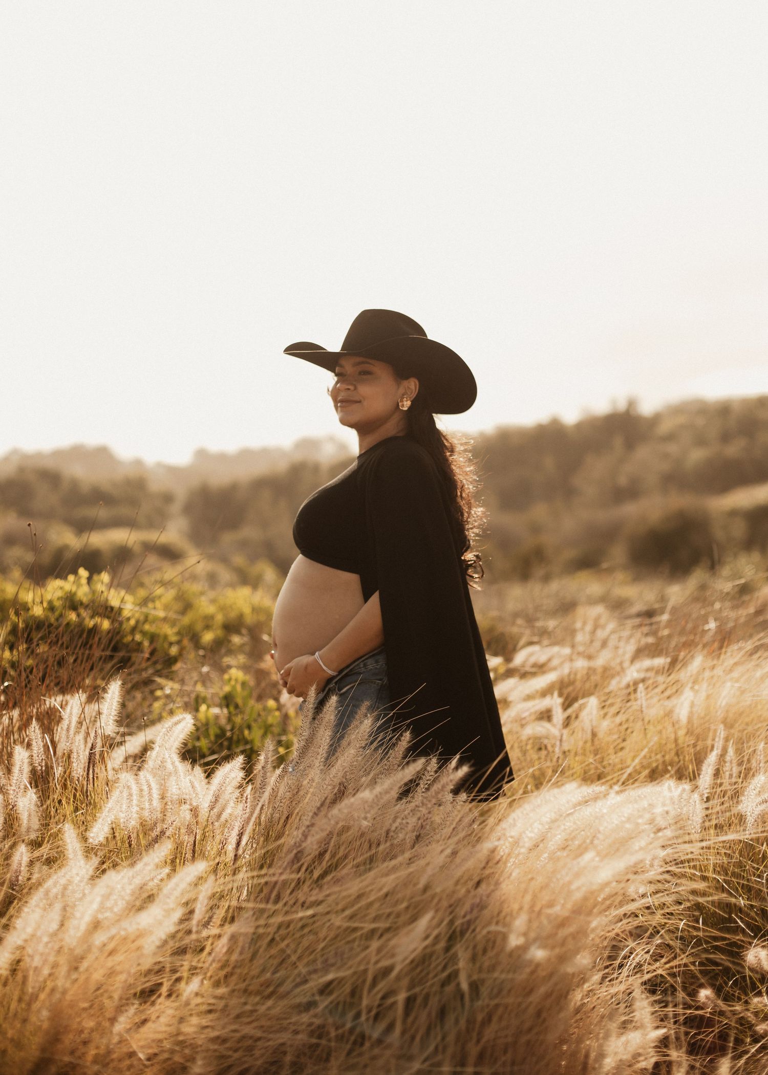 Silhouette of a pregnant woman in a cowboy hat standing in tall golden grass at sunset.