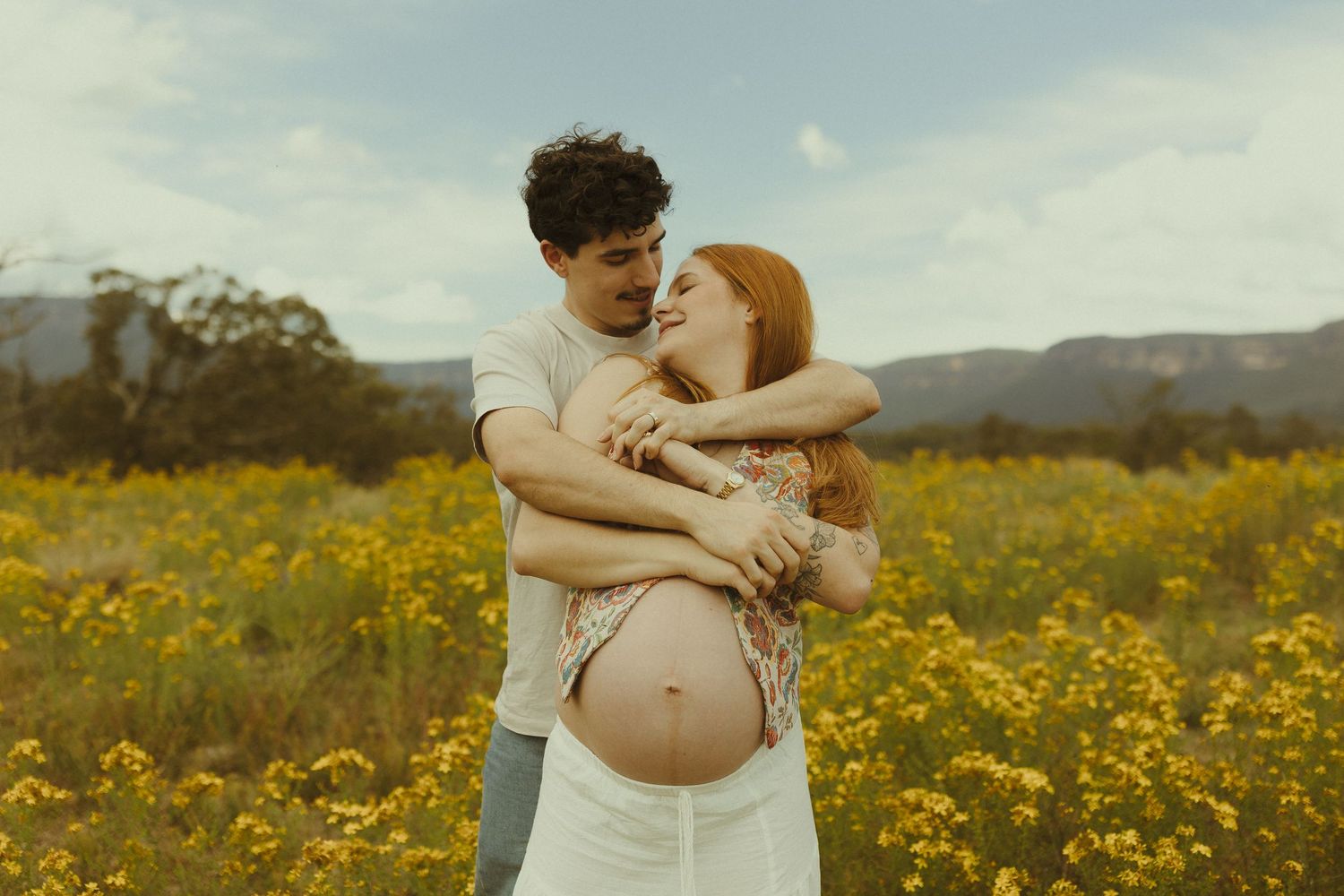 Two people share an intimate moment among blooming yellow wildflowers in a rural countryside setting.