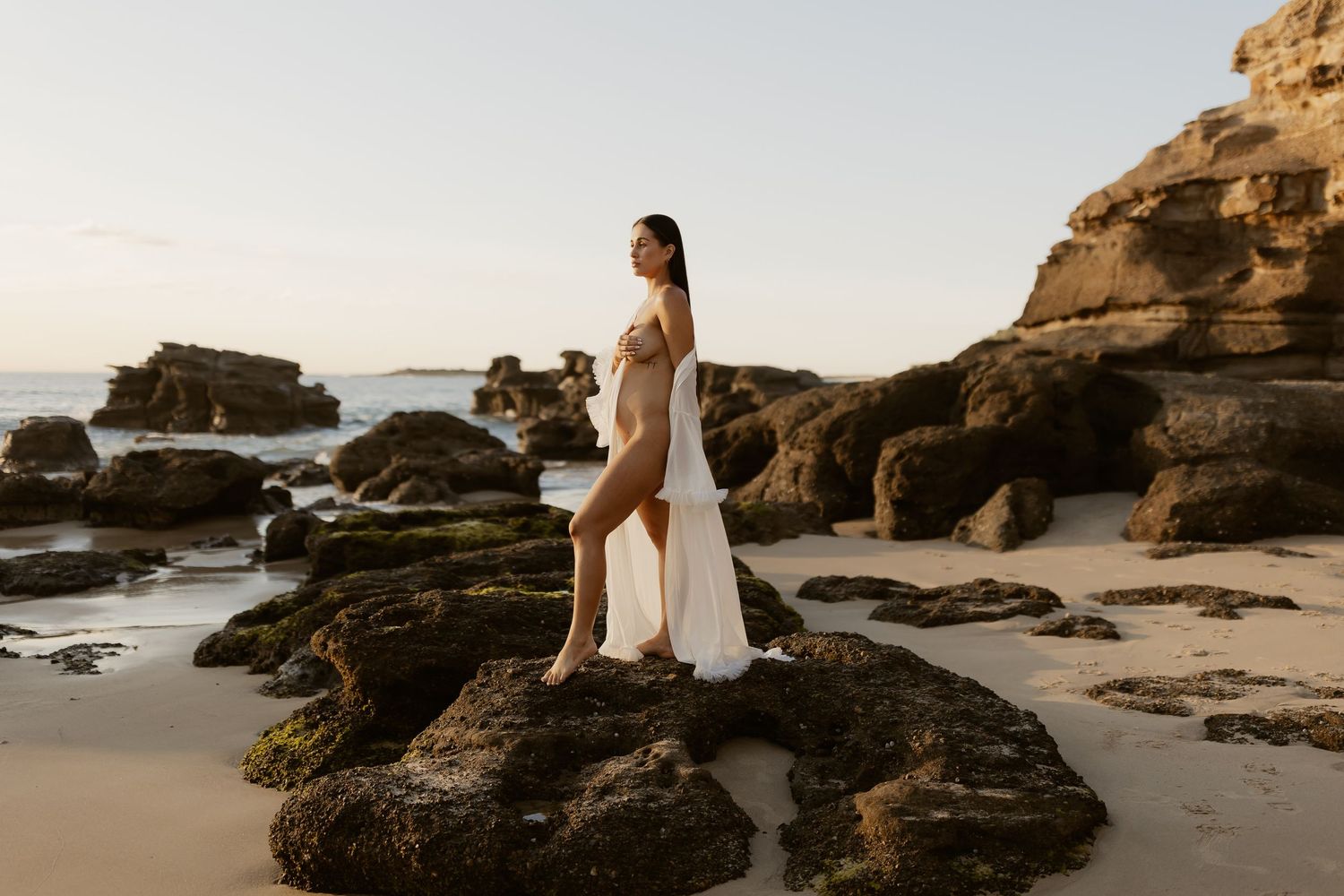 A romantic coastal photoshoot with a couple standing among dramatic rock formations at sunset on a sandy beach.