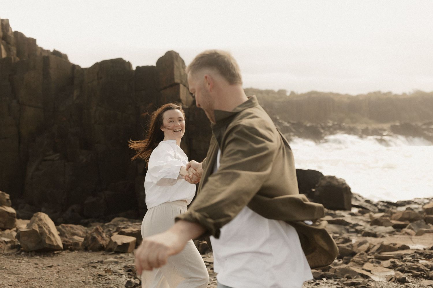 Playful couple laughs together while standing on rugged coastal rocks with ocean spray behind them.