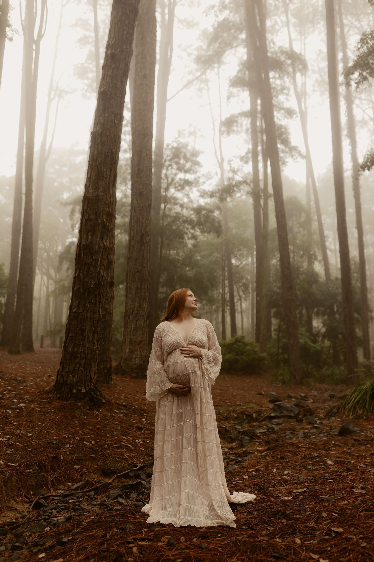 Ethereal figure in white dress stands in misty forest with tall trees creating moody atmospheric scene.