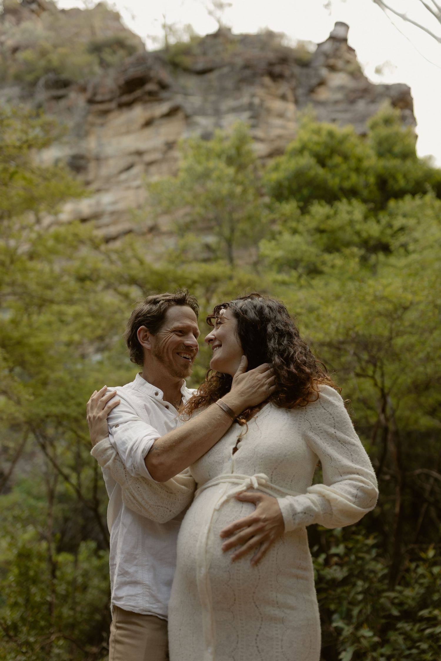 A romantic couple embraces and laughs together in front of a rocky hillside surrounded by green foliage.