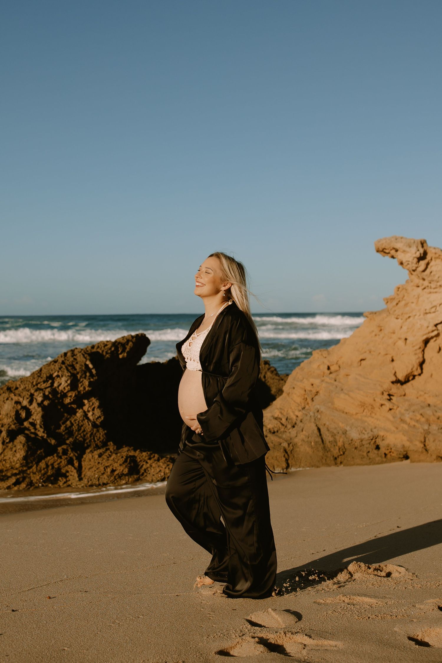 A pregnant person in dark clothing poses against a sunset beach backdrop surrounded by coastal rocks.