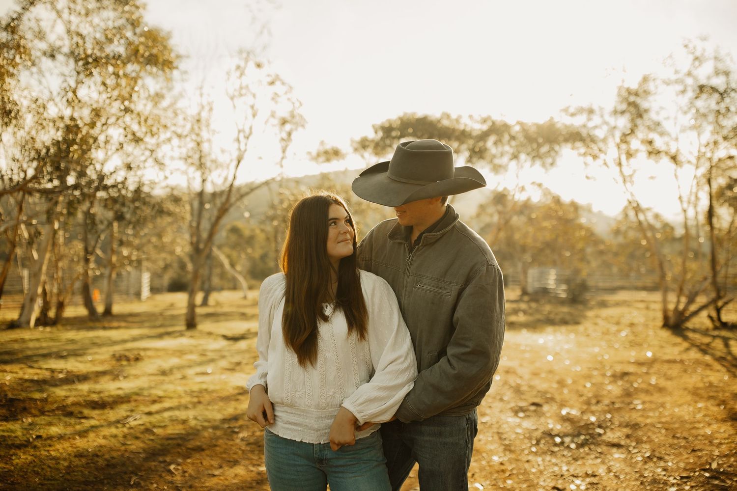 A couple embraces in a golden autumn field at sunset wearing casual clothing and a cowboy hat.
