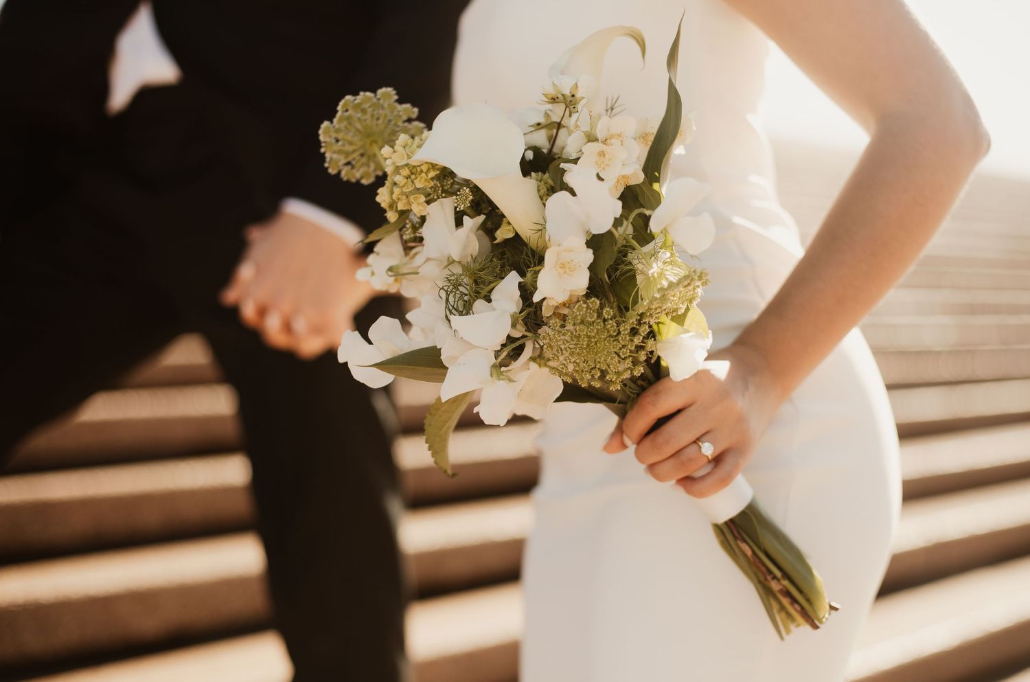 Sunlit wedding scene with bride in sleek white dress holding bouquet of white and yellow flowers.