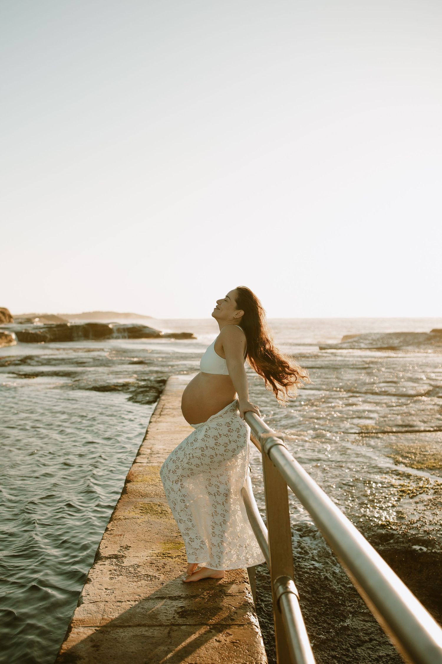 Pregnant woman in flowing dress leans on railing by ocean during sunset maternity session.