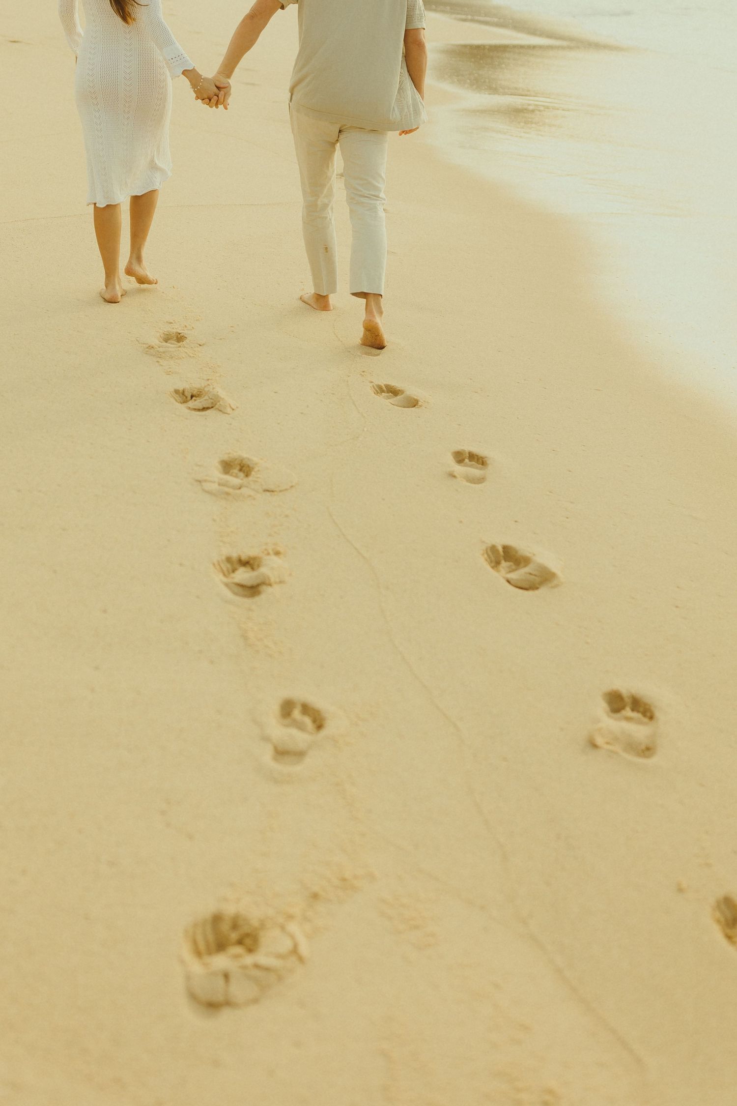 Two people walk along a sandy beach leaving footprints behind them in the golden sand at sunset.