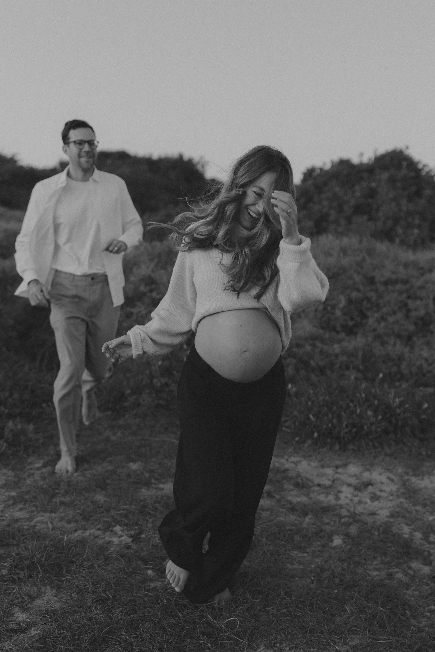 Black and white maternity photo of laughing pregnant woman with flowing hair and man in white shirt outdoors.