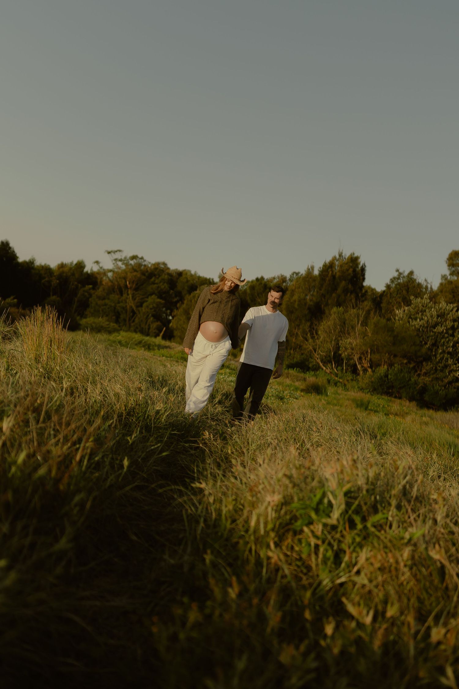 Couple walking through lush green meadow at golden hour, surrounded by trees and tall grass.