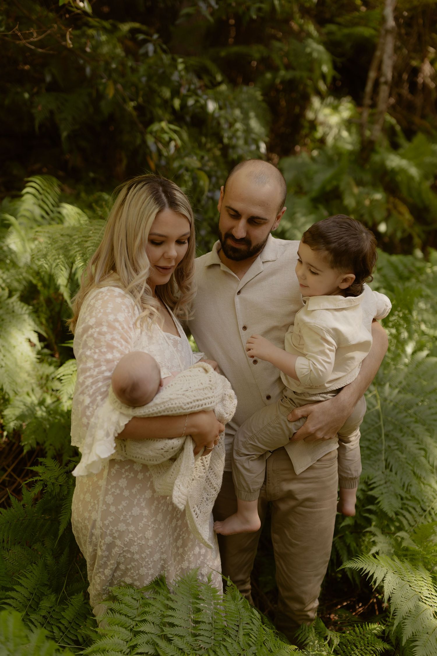 Family of four with newborn and toddler posing in green fern forest, wearing neutral tones.
