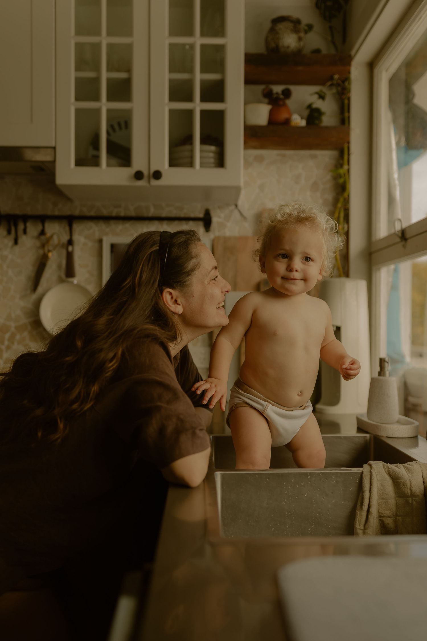 A smiling mother watches her happy toddler standing in a kitchen sink, in a warm, cozy home setting.