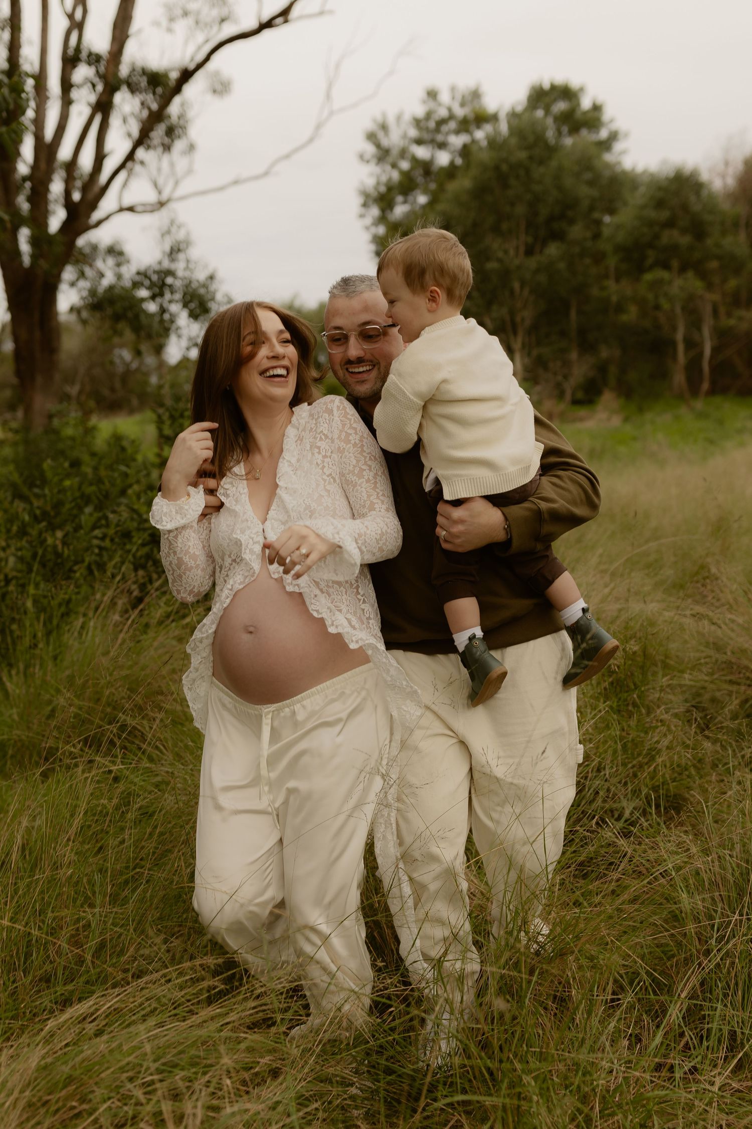 Laughing pregnant woman in lace top with partner holding toddler in lush green field.