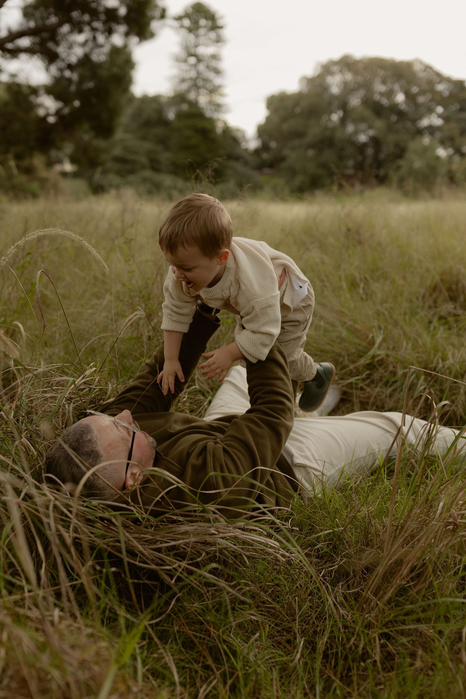 Father lies in tall grass while energetic toddler in cream sweater playfully climbs on him.