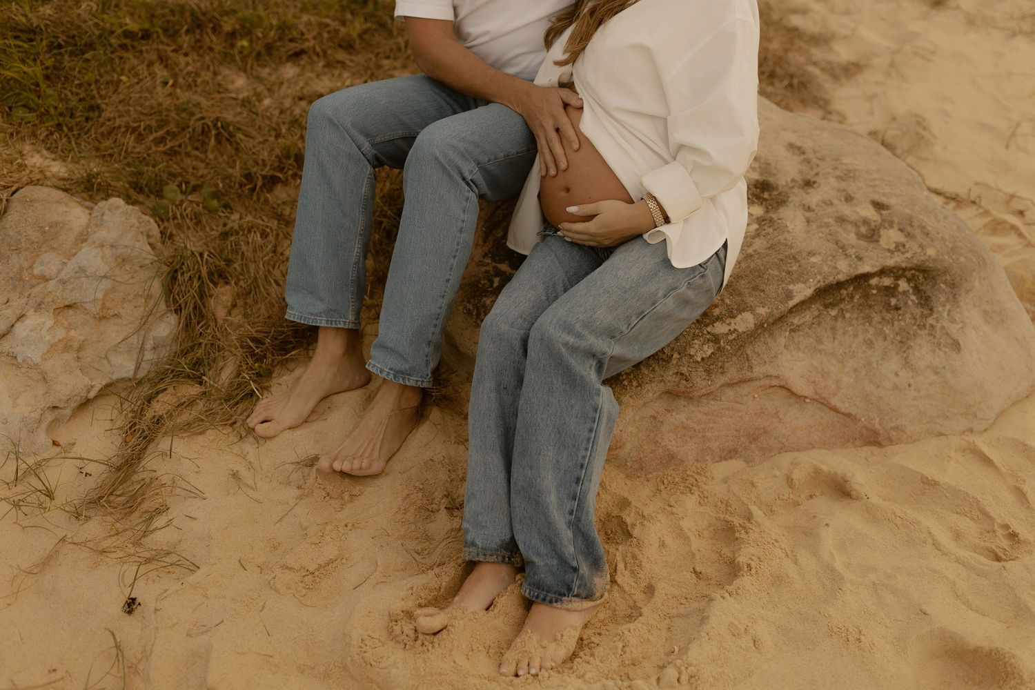 Barefoot couple sits on sandy beach rocks, partners hand resting on pregnant belly, casual jeans.