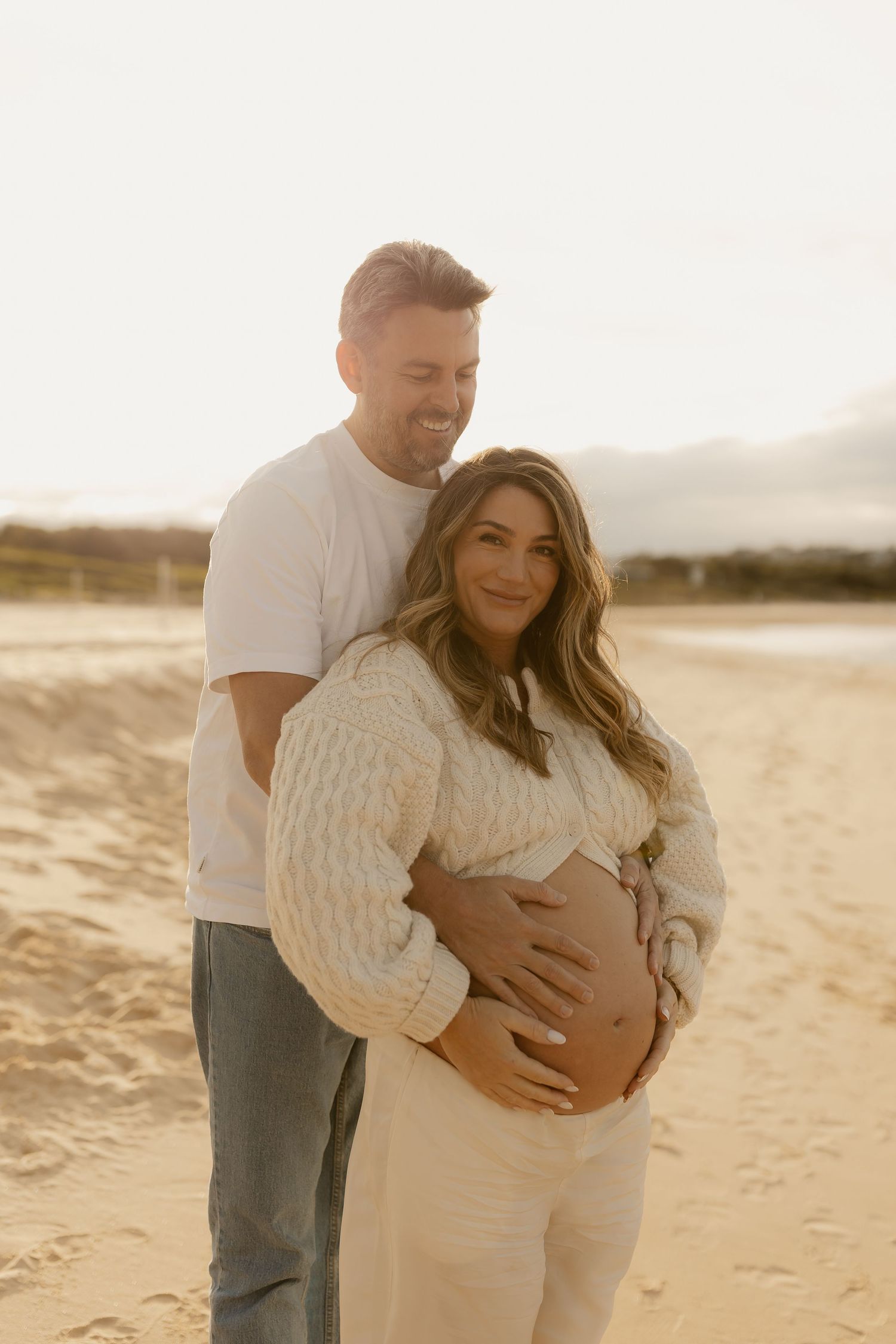Couple poses on sunlit beach, man embraces pregnant woman from behind, both smiling warmly.