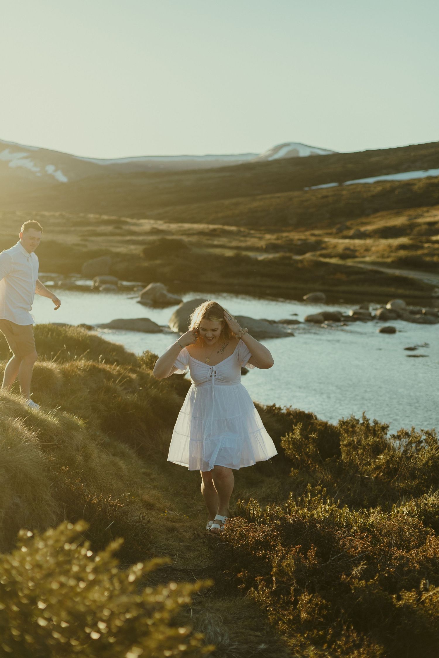 Couple walking along coastal trail during golden sunset with mountains and water in background.