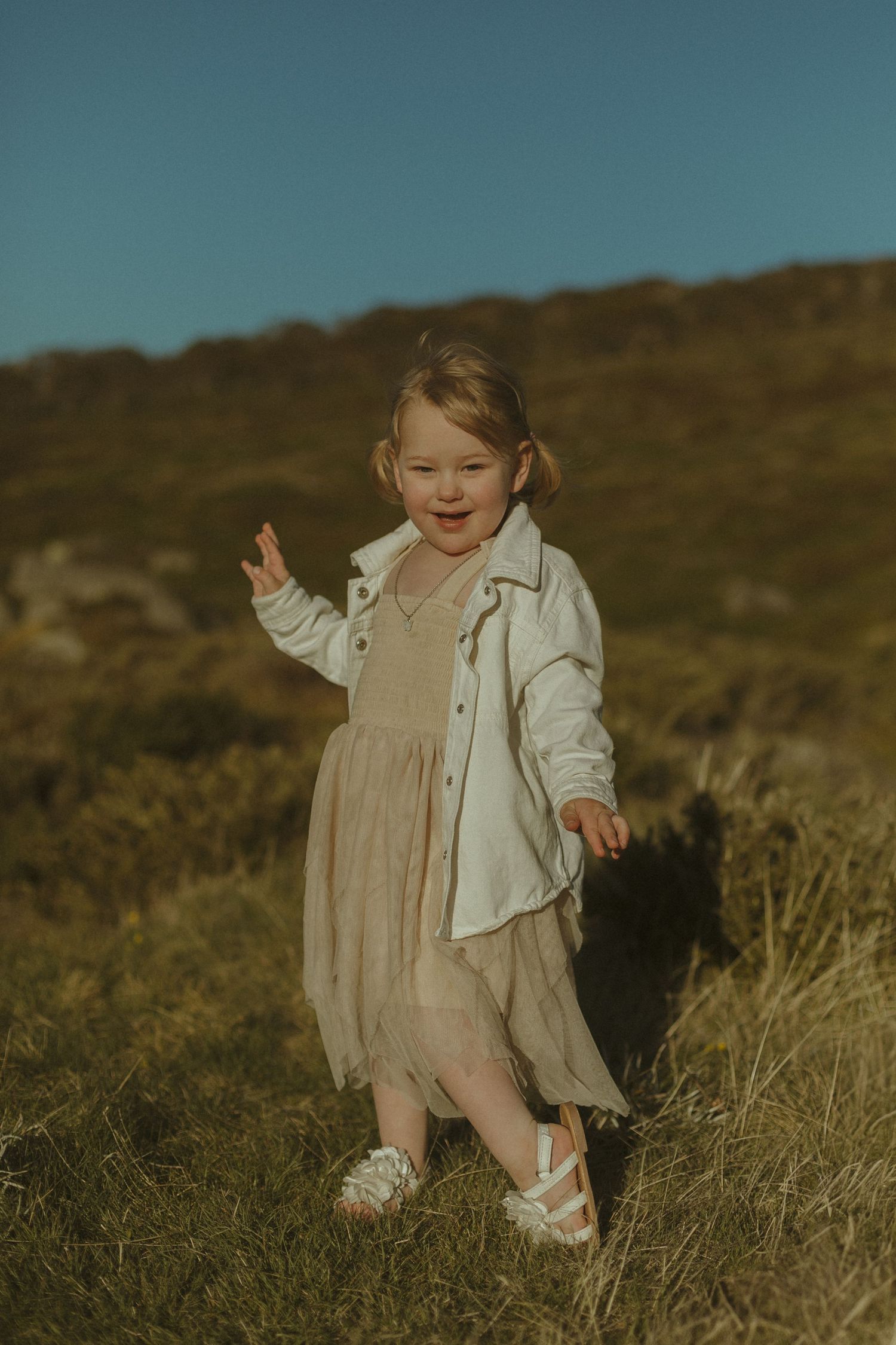 A small child wearing a white jacket and dress stands in a grassy field during golden hour, smiling with joy.