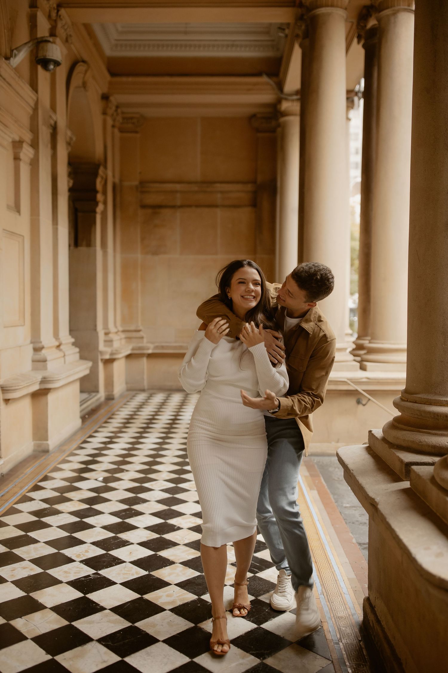 A romantic couple shares a tender moment in an elegant hallway with checkerboard floors and classical columns.