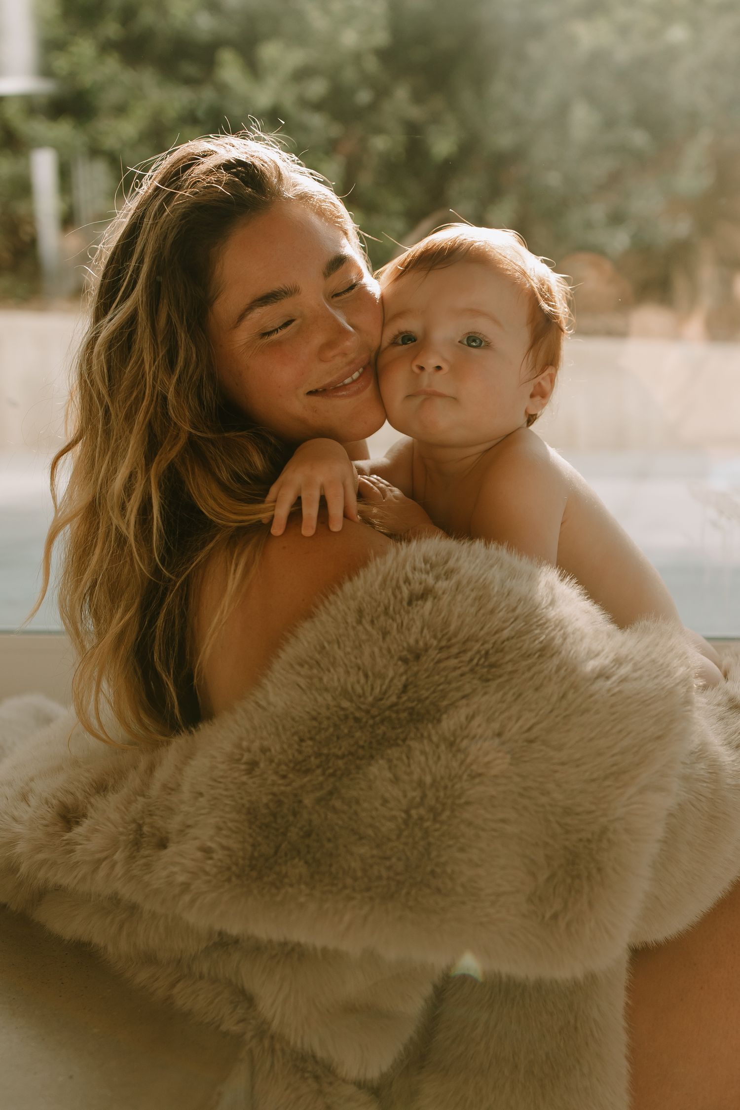 Intimate portrait of parent and baby wrapped in soft fuzzy blanket during tender golden hour moment.