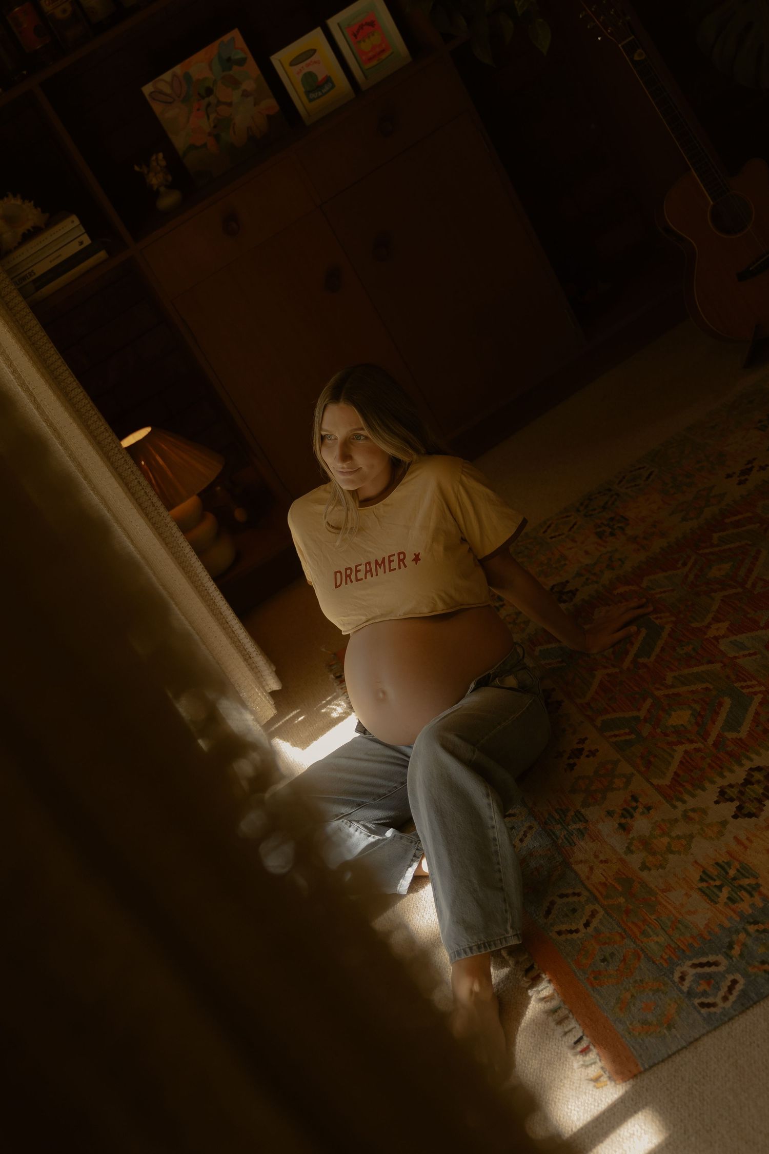 Pregnant woman in a Dreamer shirt sits on floor in warm light, cradling her bare baby bump.
