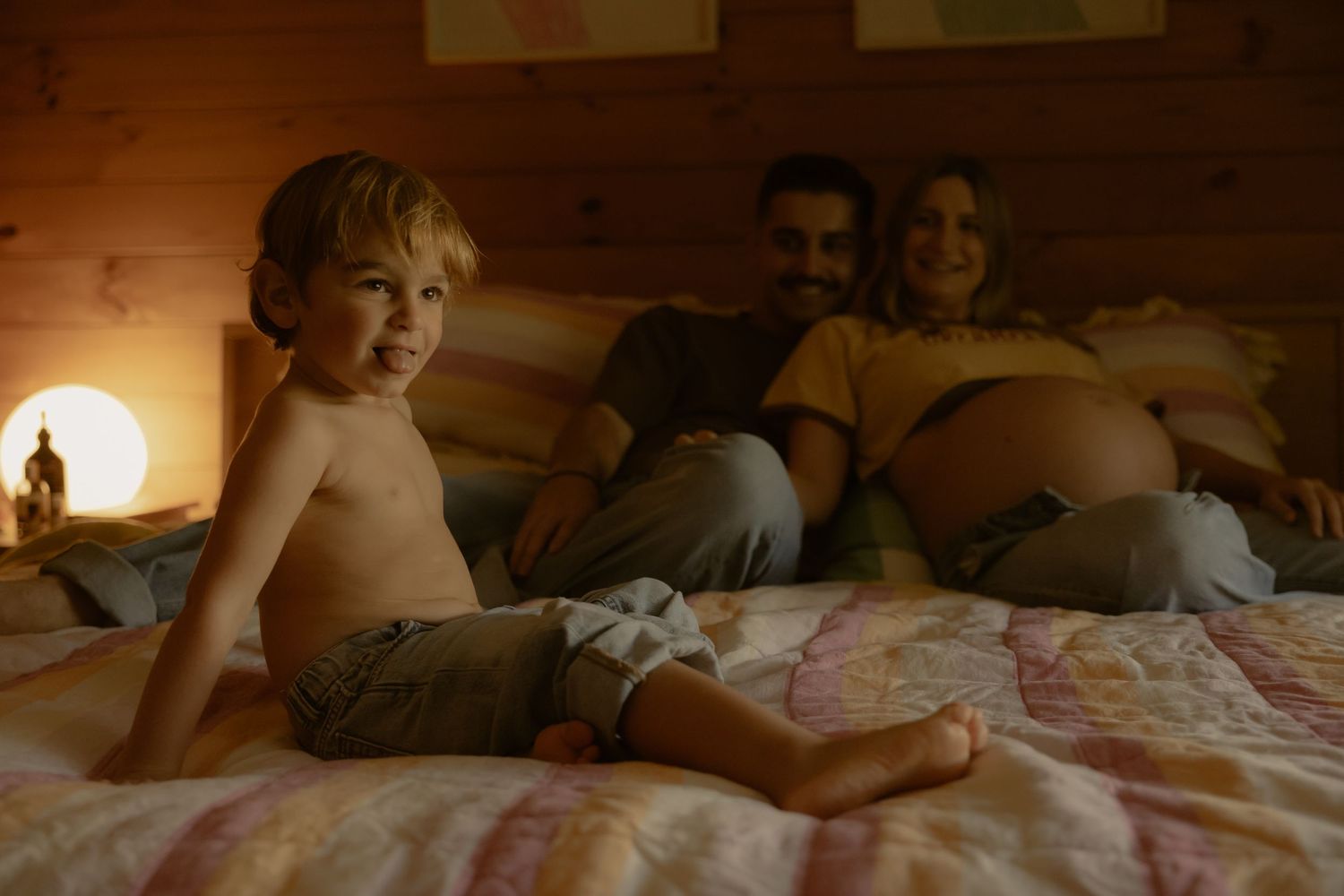 A smiling toddler sits on a bed with his parents relaxing behind him in a cozy lit bedroom.