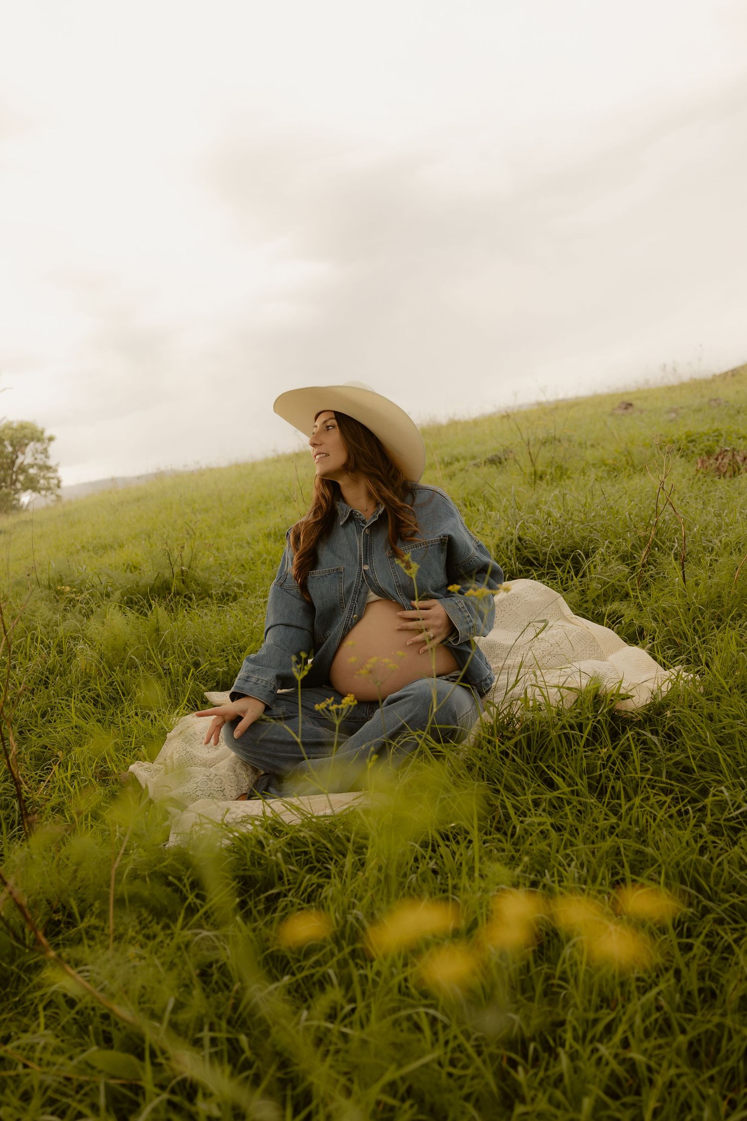 Pregnant woman in denim and wide-brim hat sits on green meadow, cradling her baby bump while looking up dreamily.