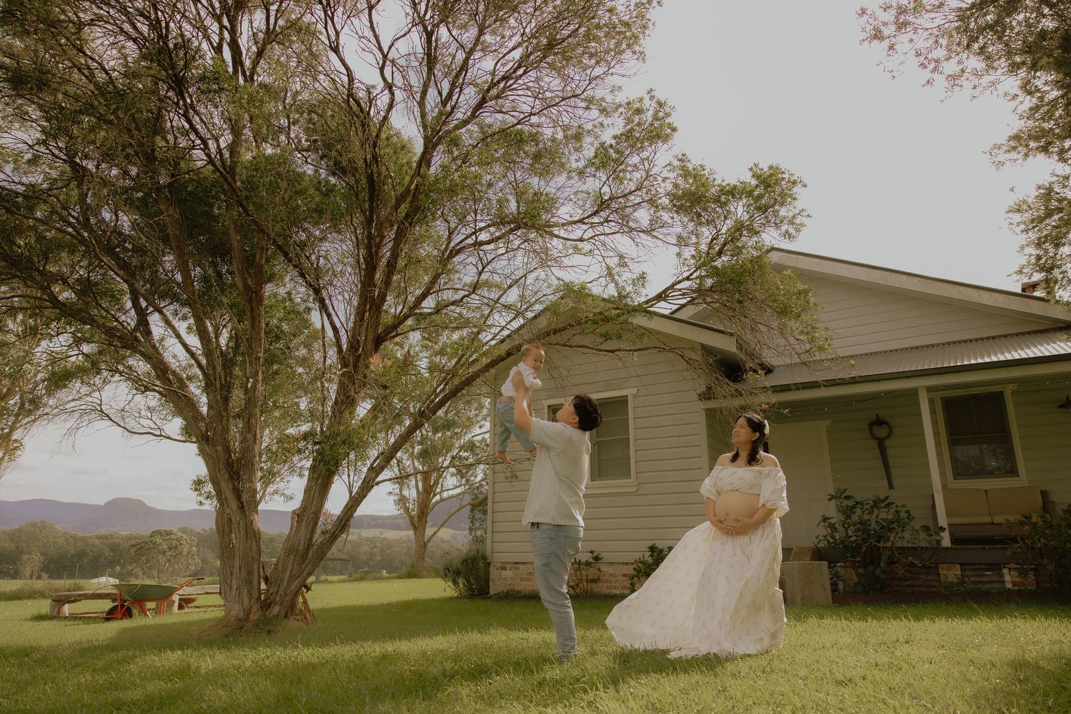 Pregnant woman in white dress dances with partner outdoors near large tree beside rural farmhouse on green lawn.