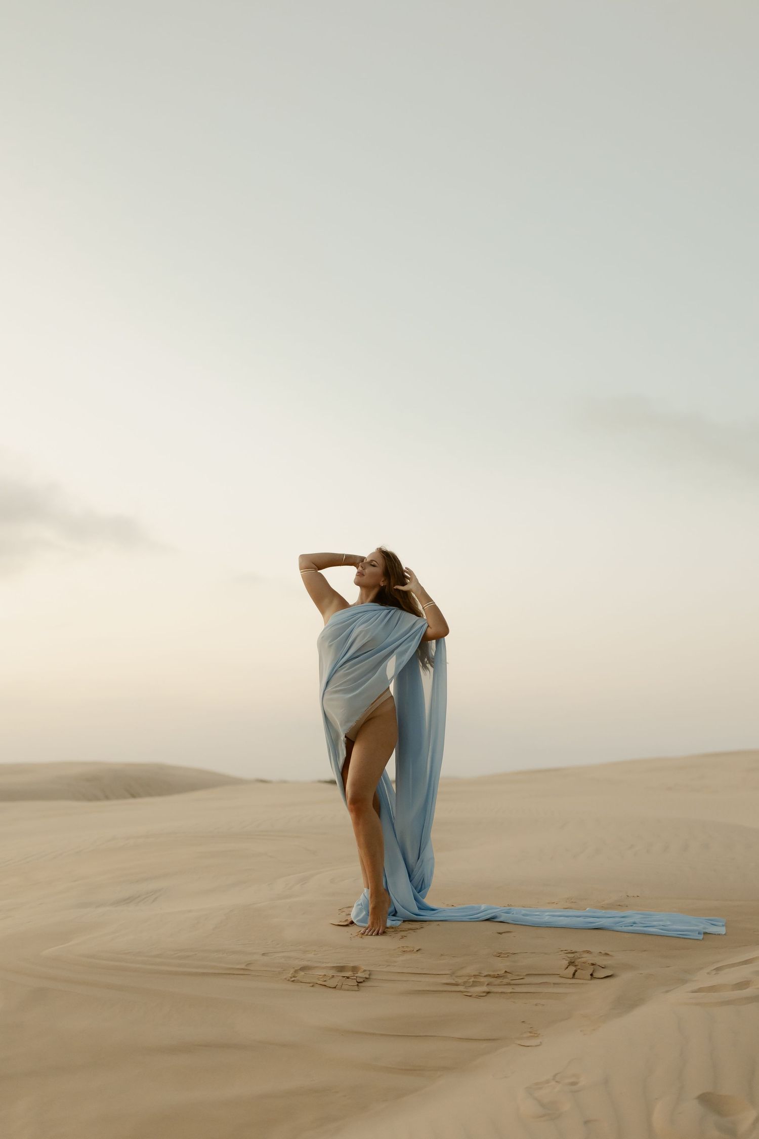 A person wrapped in flowing blue fabric poses gracefully across desert sand dunes at sunset.