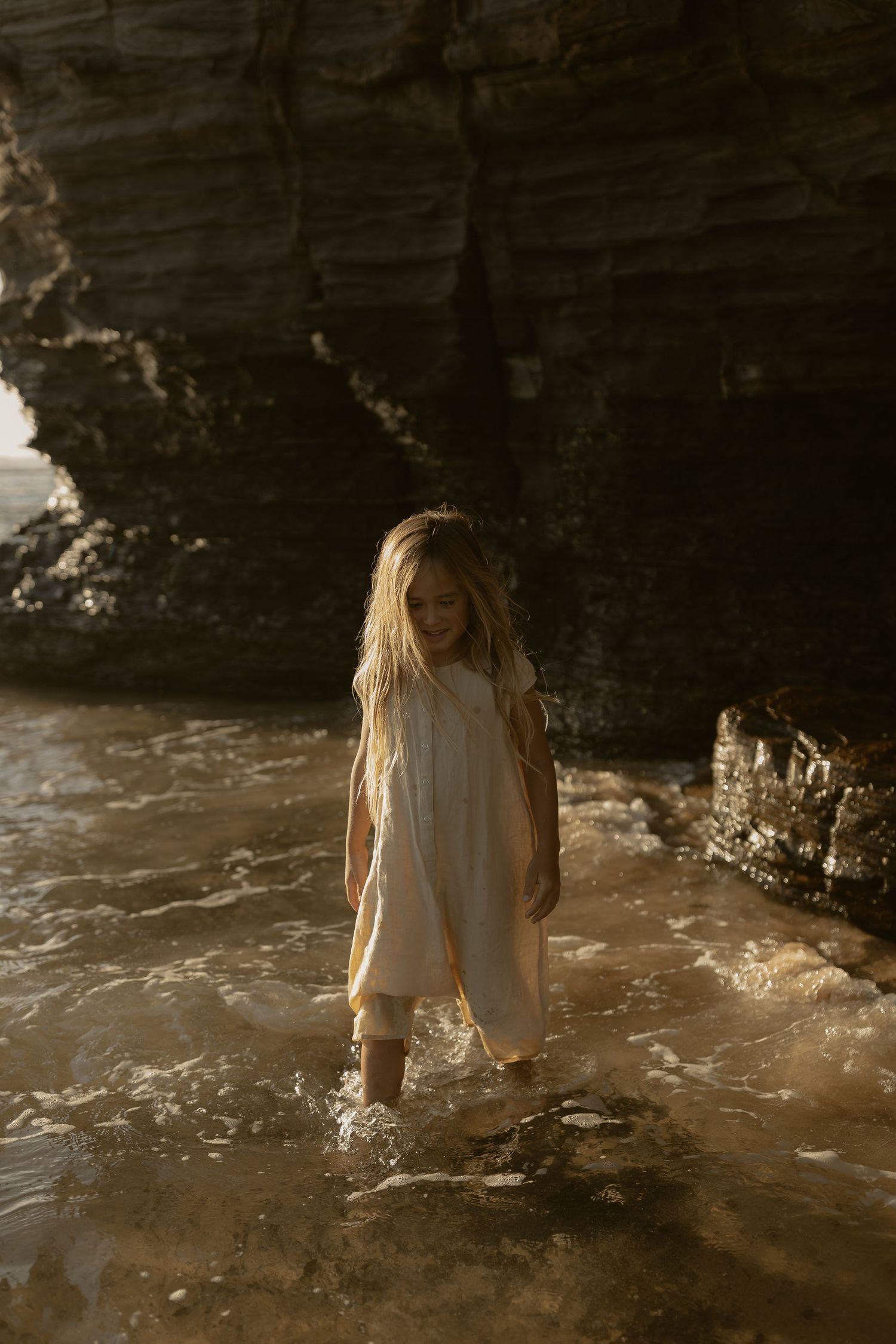 A figure in a white dress stands near rocky cliffs by the ocean at sunset, creating a dreamy coastal atmosphere.