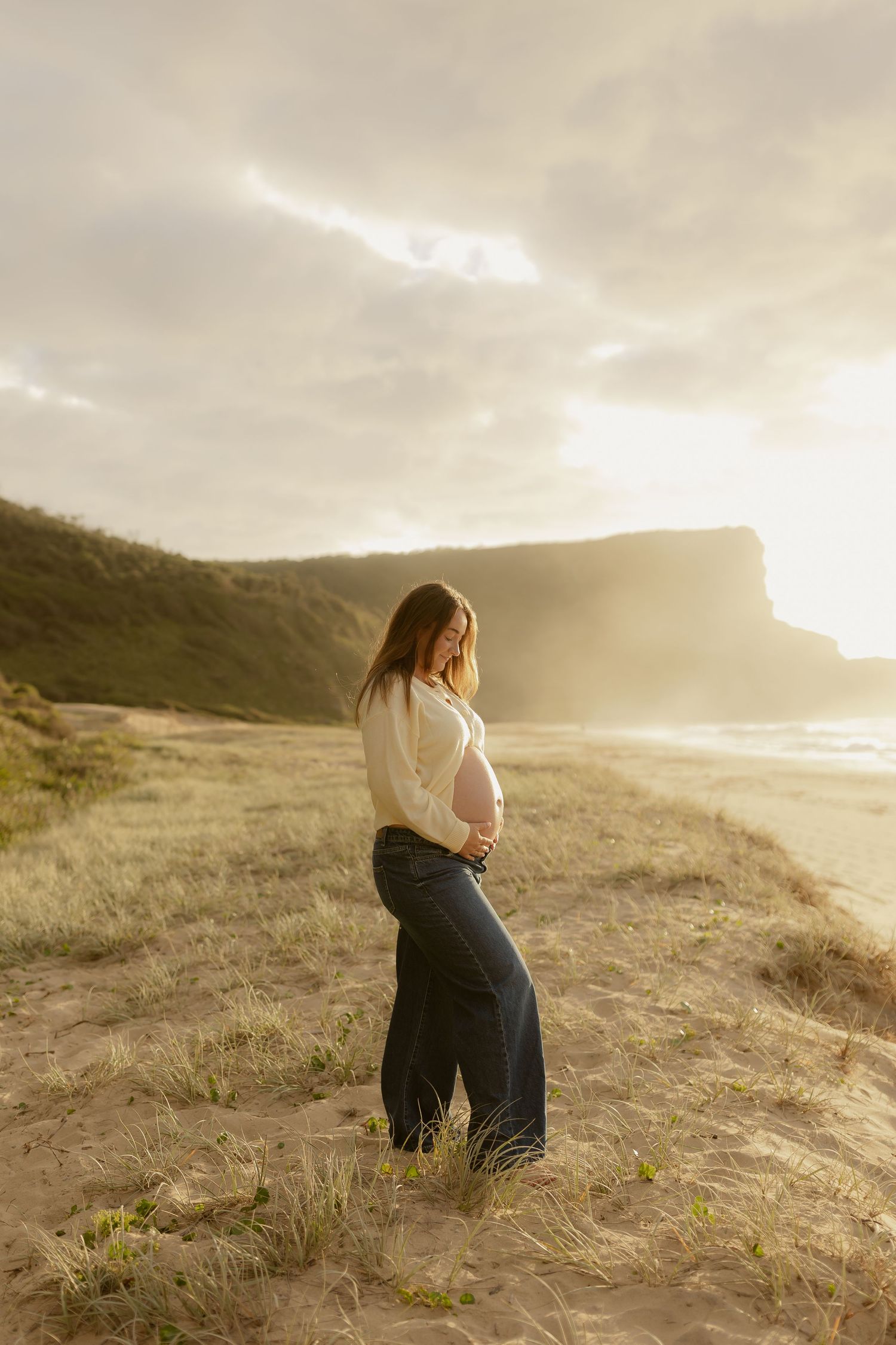A person in a white sweater sits on a grassy coastal hillside during golden hour sunset.