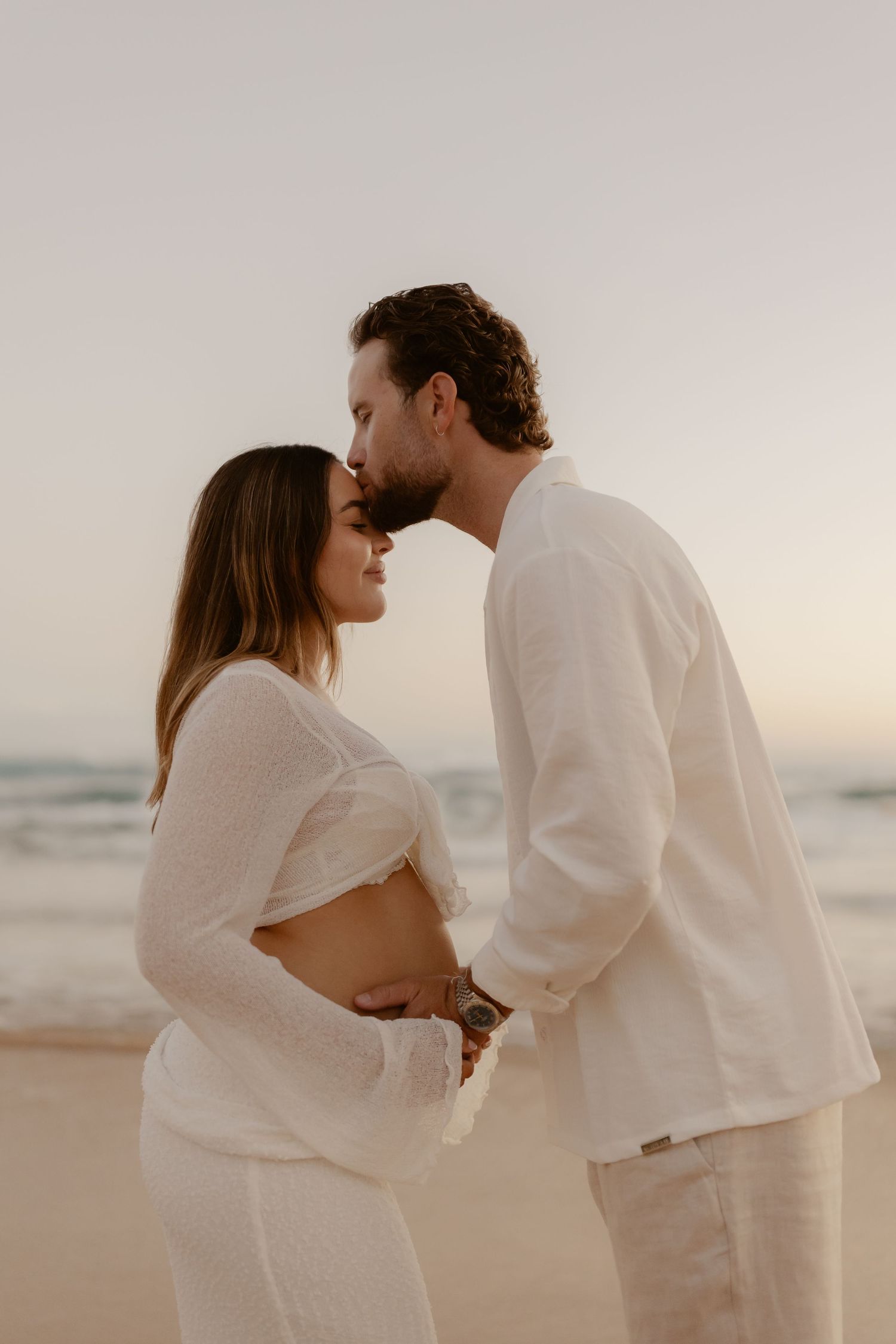 An intimate maternity photo session on the beach with a couple wearing matching white outfits.