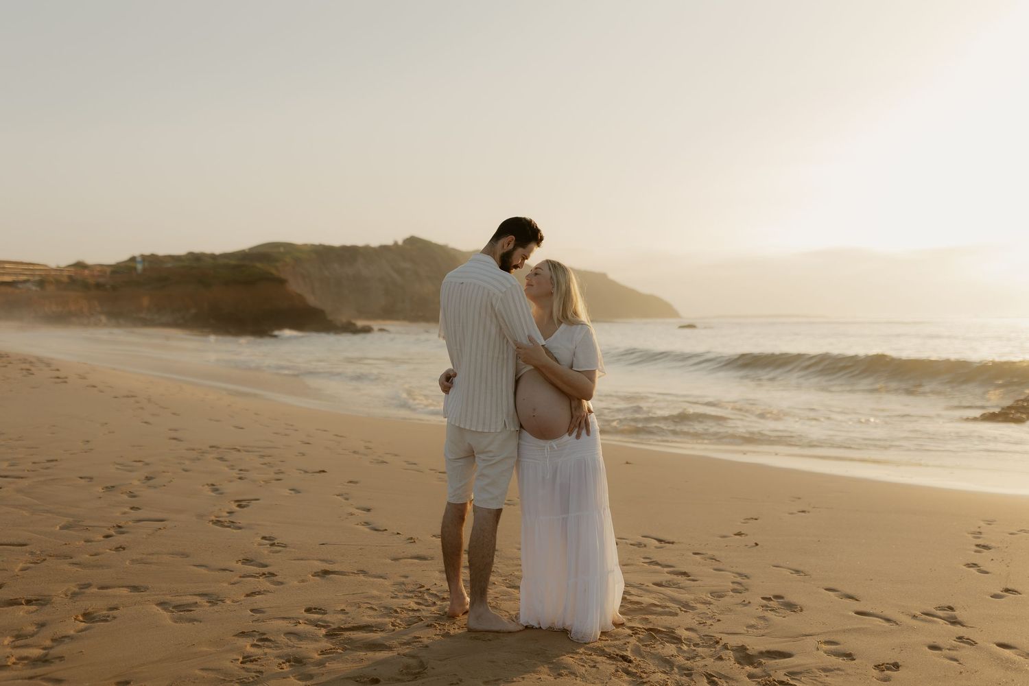 A couple embraces on a sandy beach at sunset with ocean waves and mountains in the background.