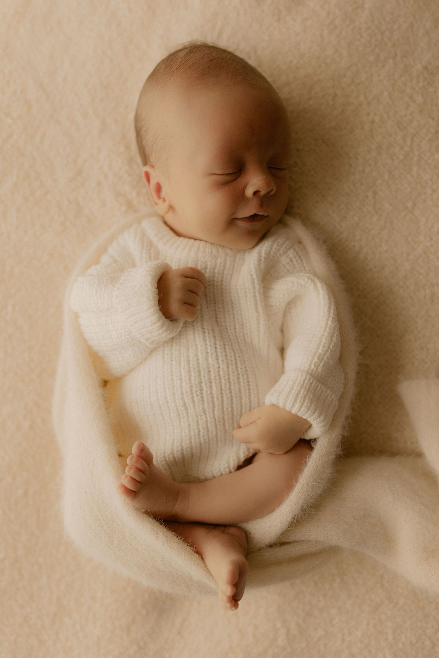 Newborn baby wearing white knit sweater lying on cream colored blanket in artistic panoramic composition.