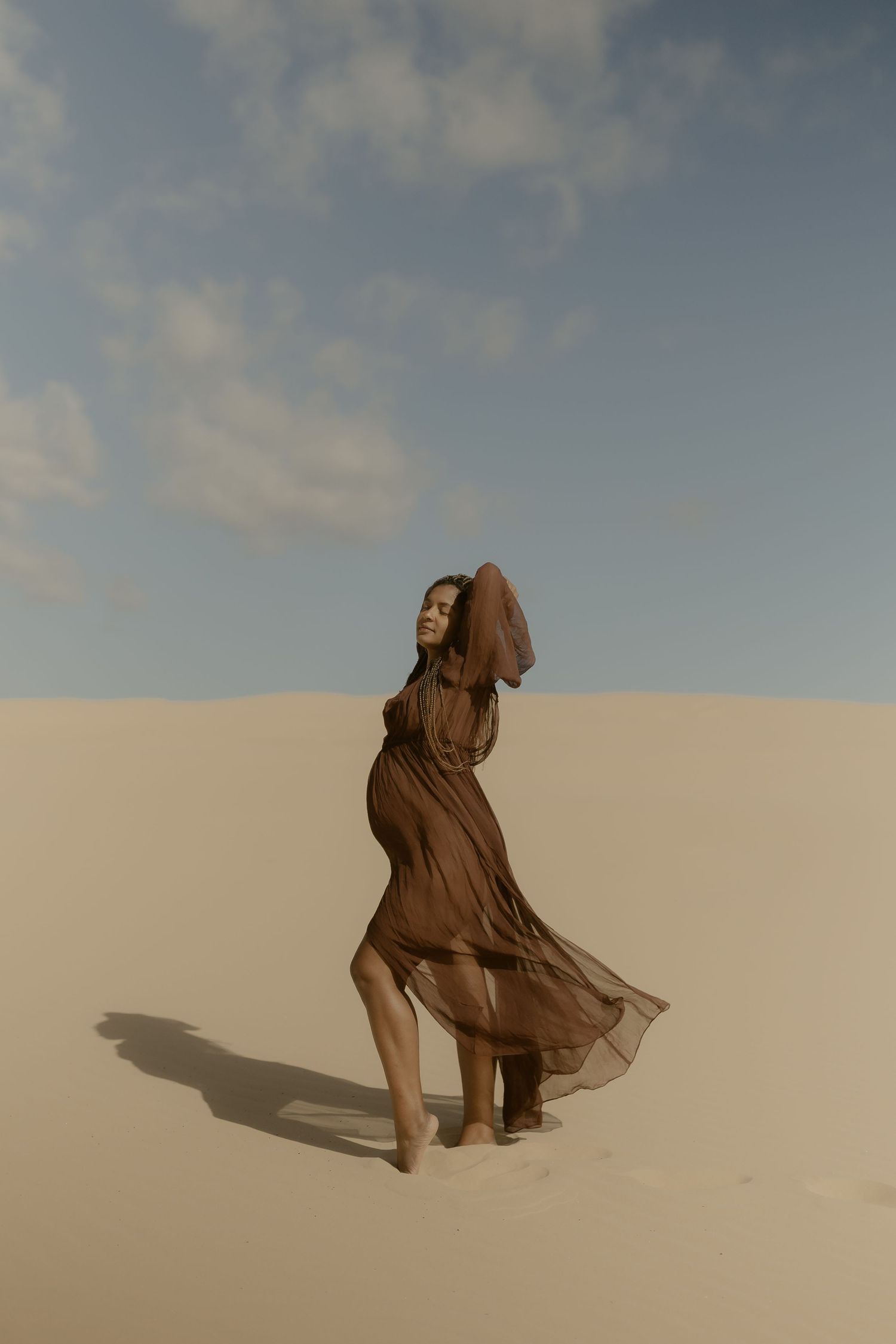 A figure in flowing brown fabric stands dramatically against sand dunes and cloudy sky in a desert landscape.