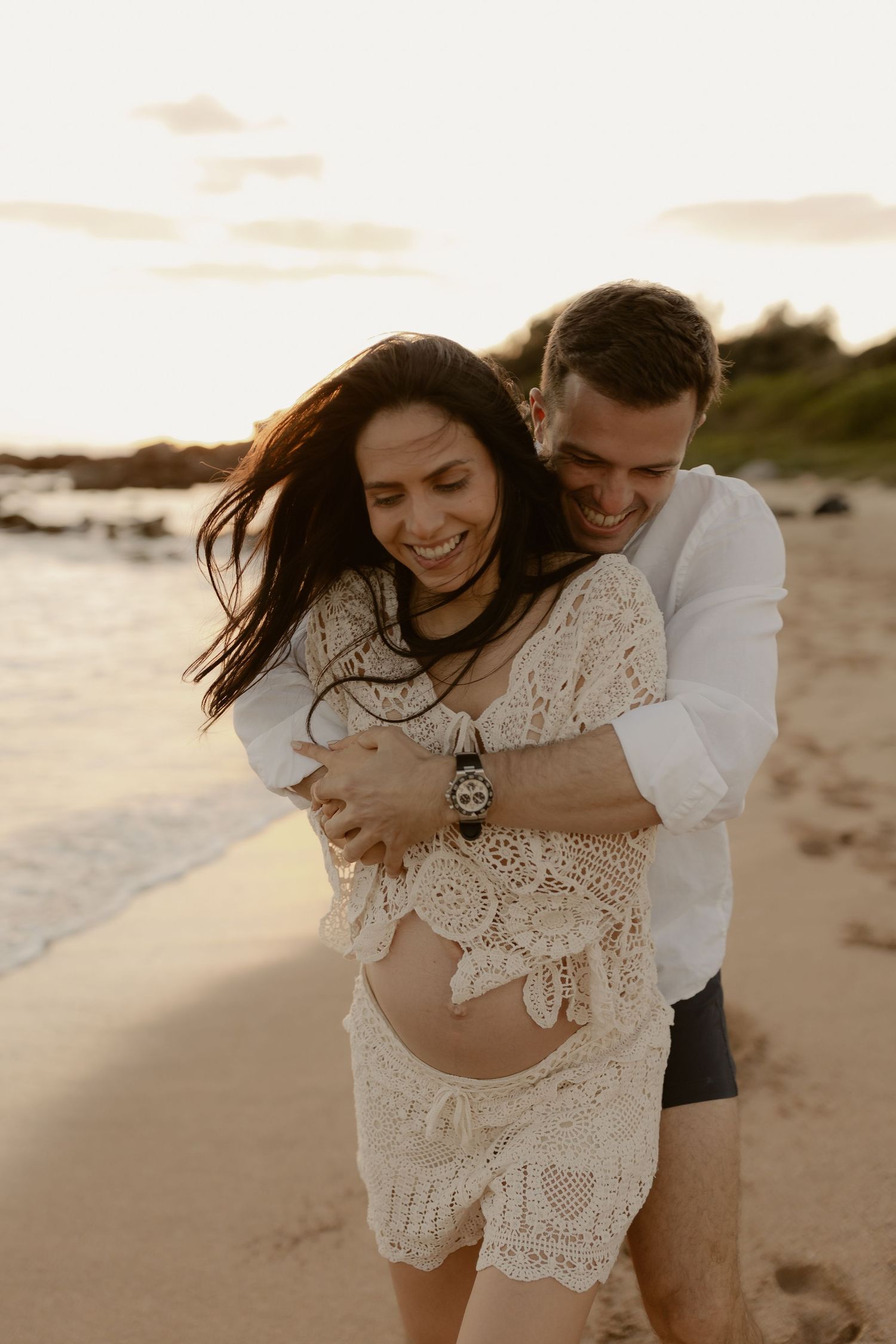A romantic couple embraces on a sandy beach at sunset, the bride wearing a flowing lace dress as her hair blows in the wind.