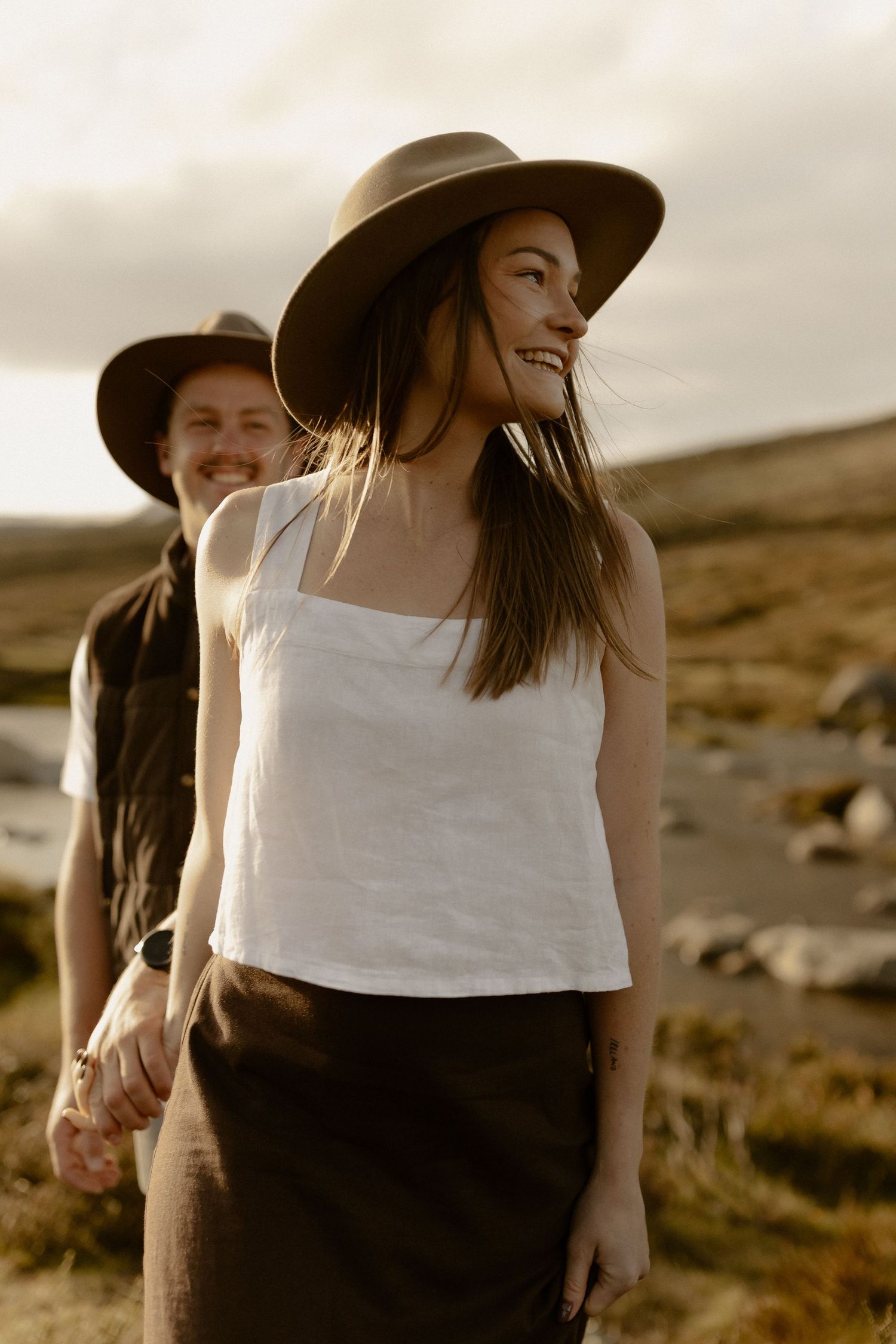 A couple in casual attire and wide-brimmed hats share a joyful moment against a desert landscape at sunset.
