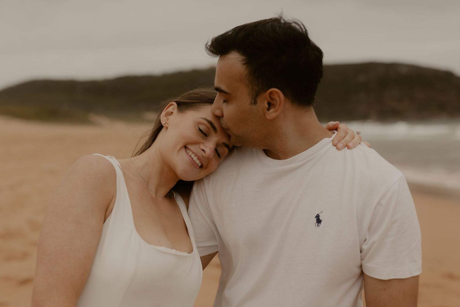 A couple embraces on a sandy beach at sunset, sharing an intimate moment in white casual attire.