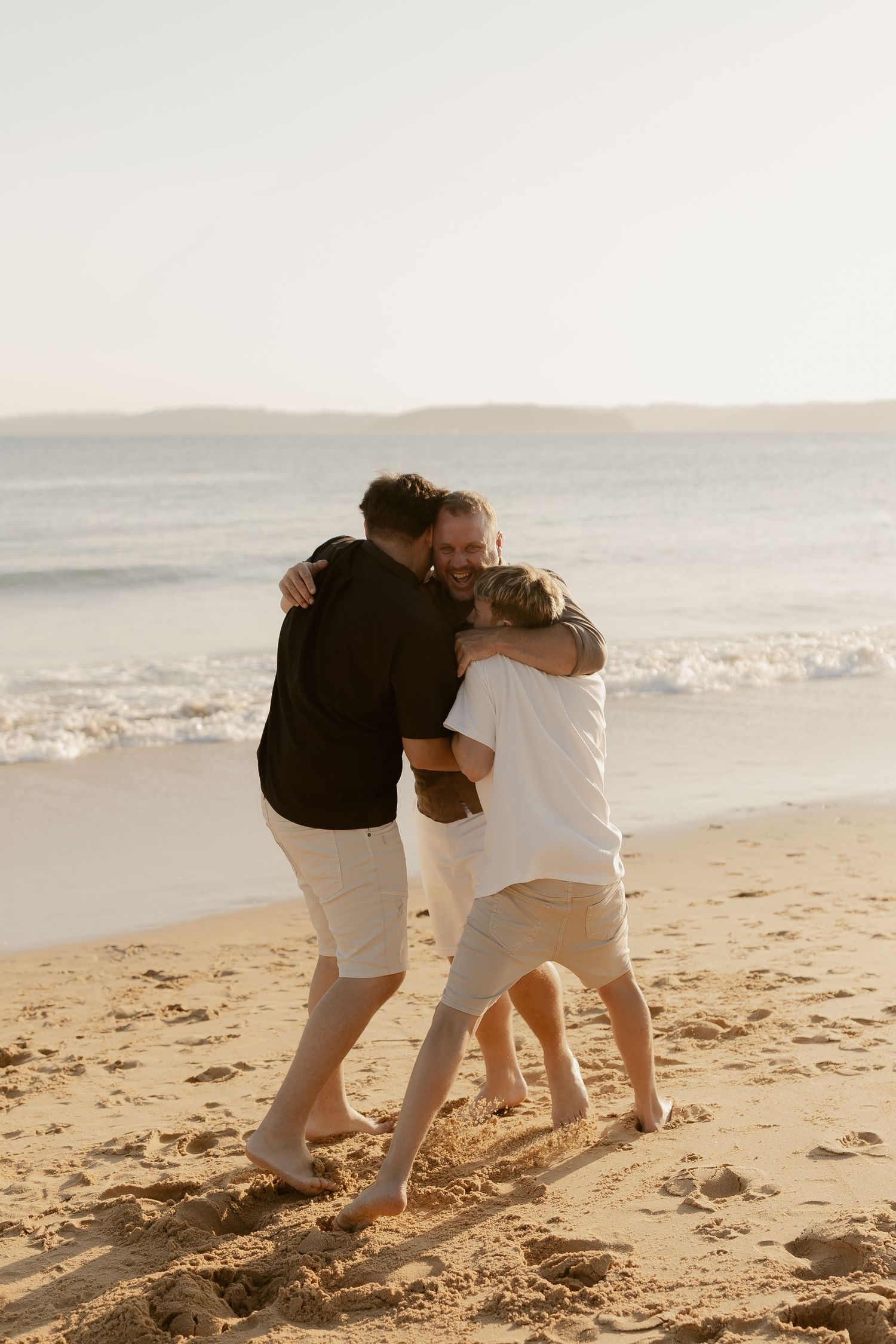 Three people embrace in a warm group hug on a sandy beach during golden hour.