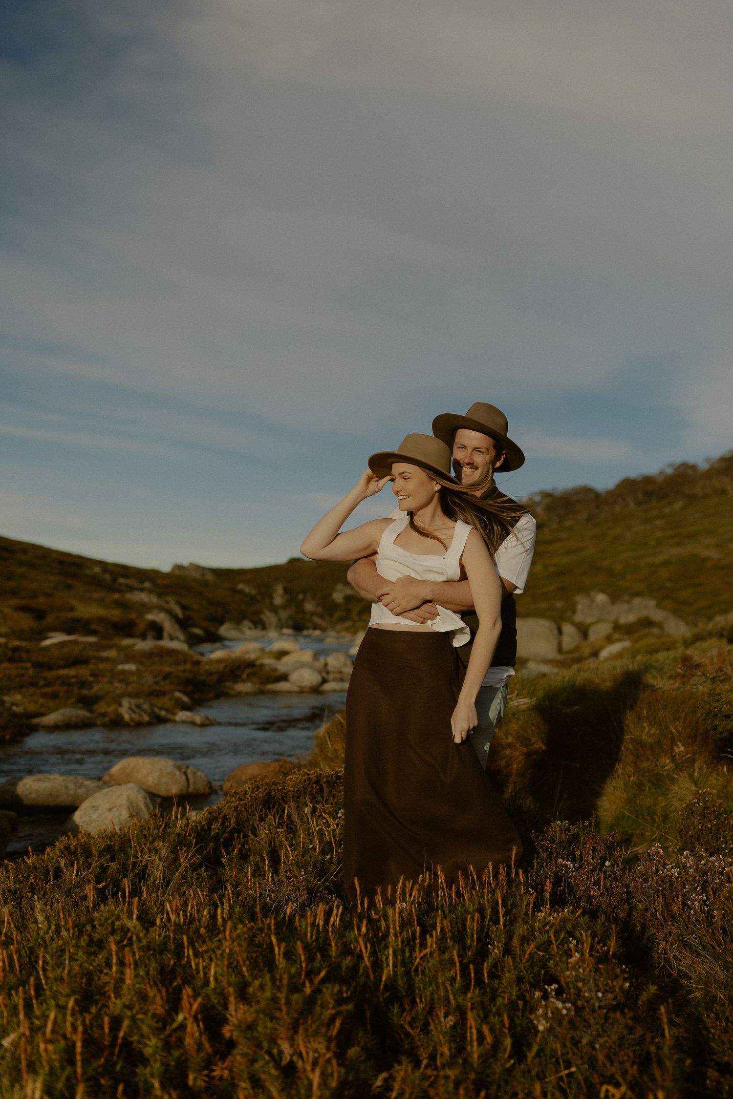 A romantic couple embraces at sunset by a stream in a rustic outdoor setting with golden light.